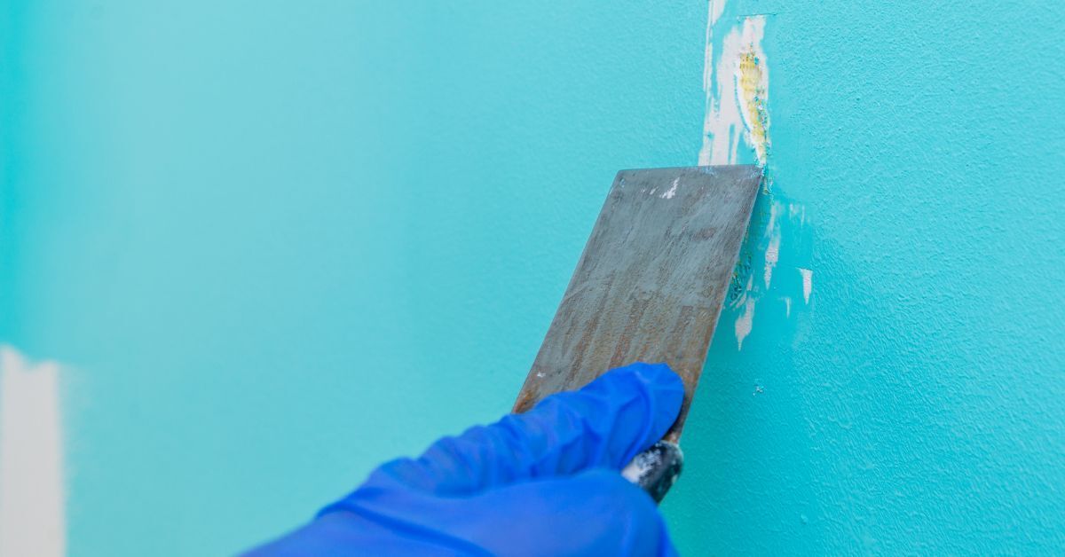 Gloved hand using a putty knife to apply spackle on a turquoise wall.