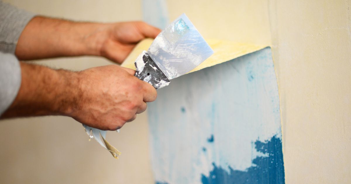 Person removing wallpaper with a scraper, light blue paint visible.