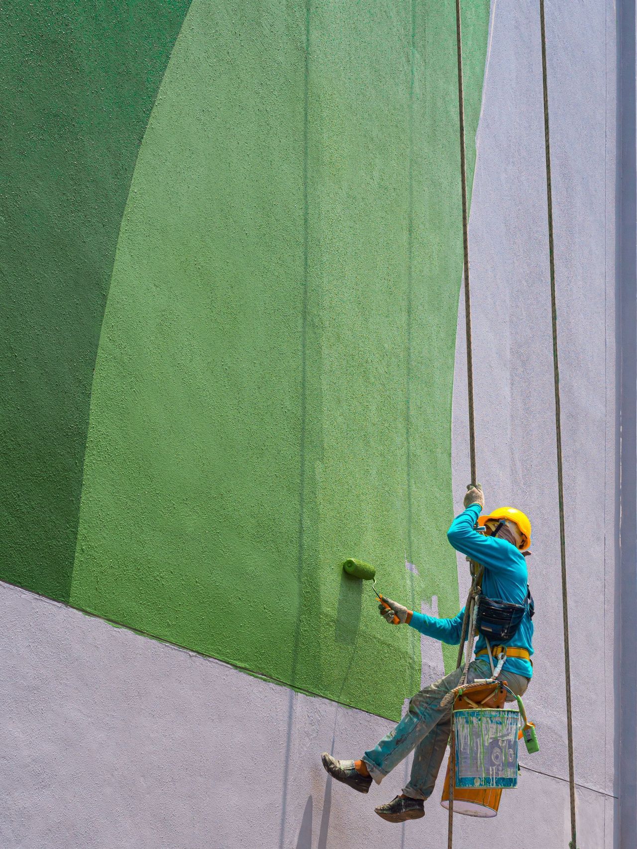 Person on ropes paints a building's wall with green paint. The building has different shades of green.