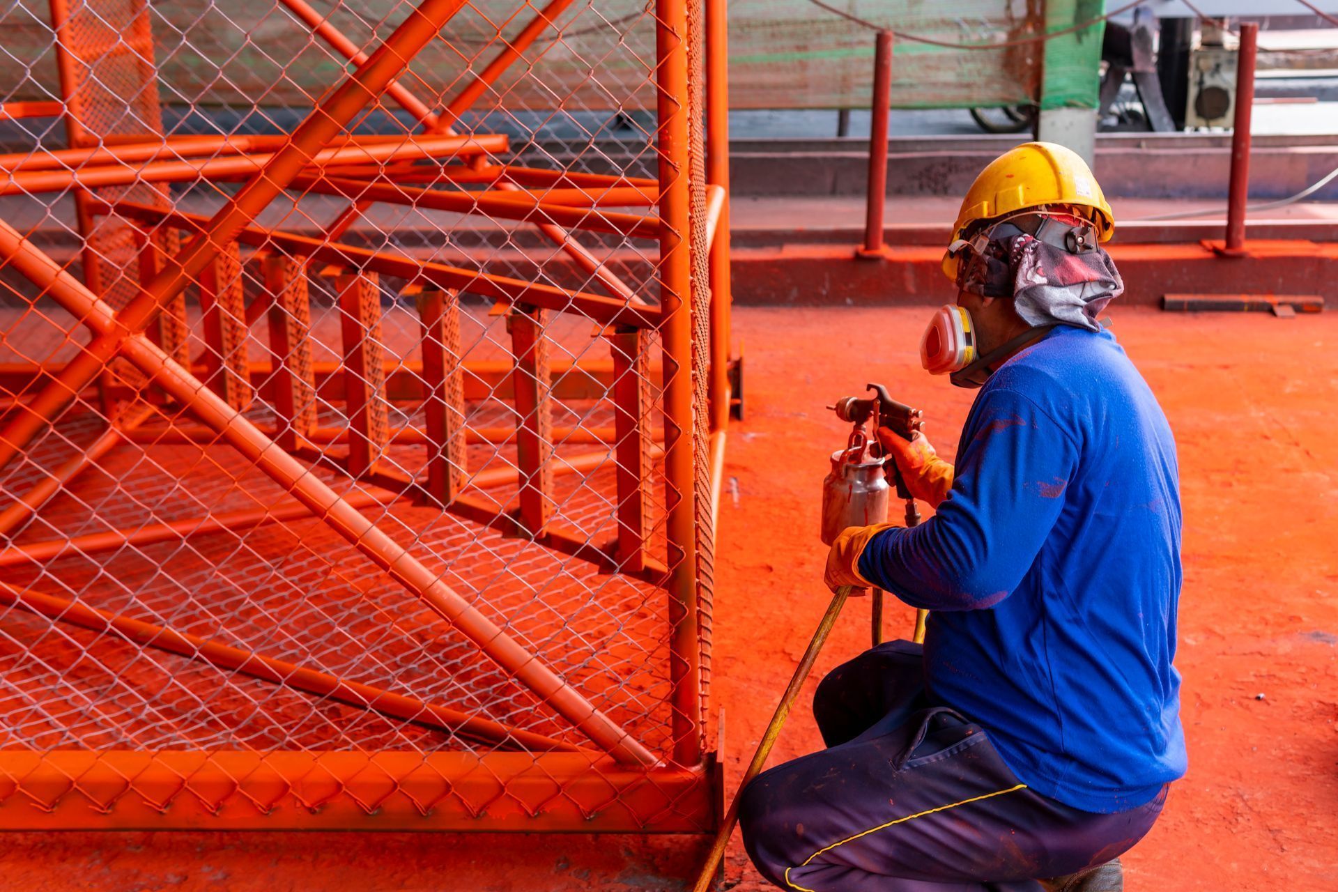 Person in safety gear spray-painting orange structure in industrial setting.