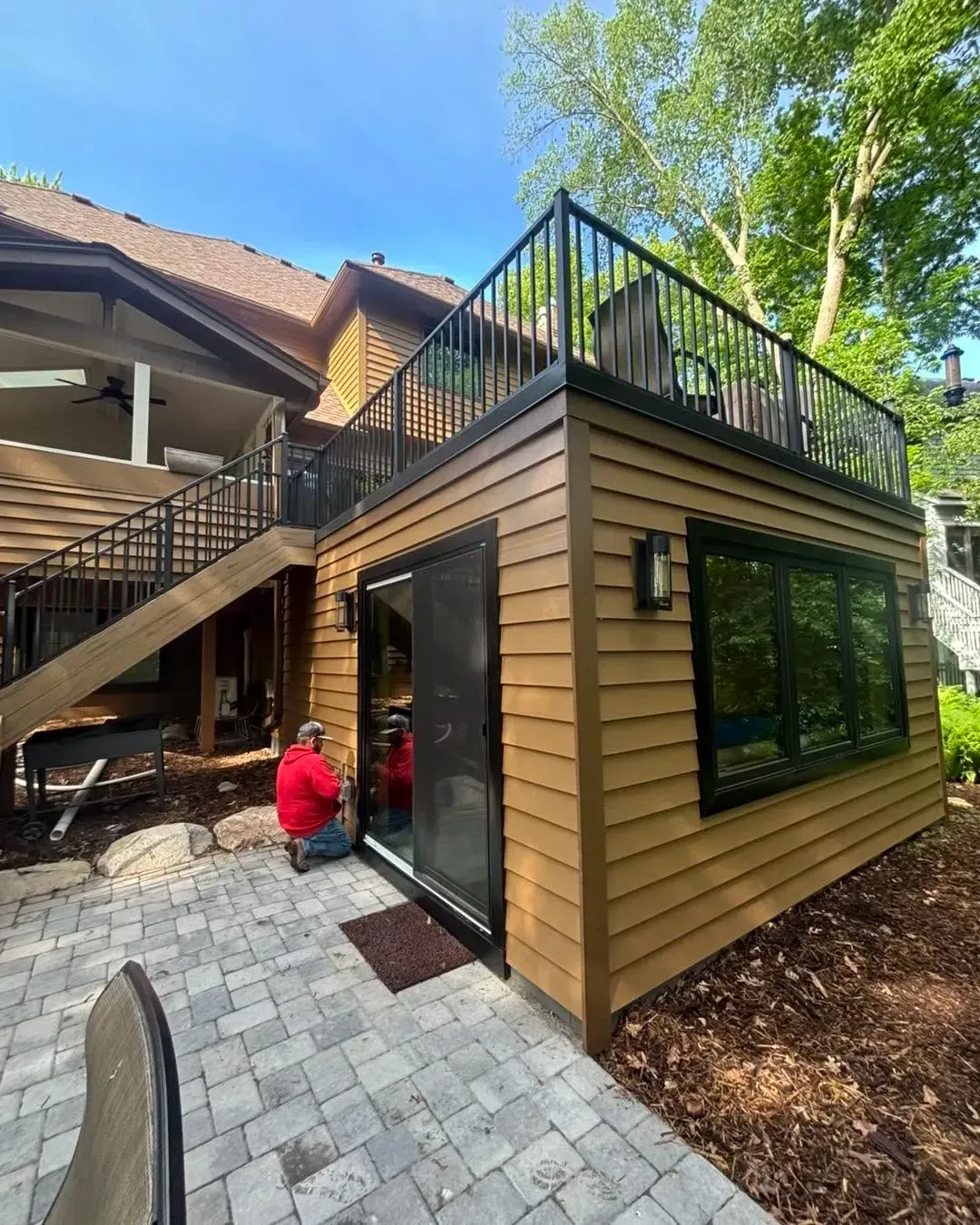 Modern tan-sided patio addition with rooftop deck, black railings, and large windows beside brick pavers Modern tan-sided patio addition with rooftop deck, black railings, and large windows beside brick pavers