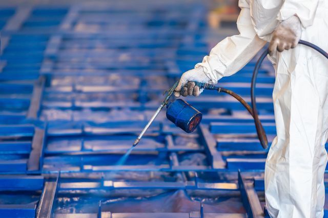 Person in protective suit sprays blue paint on metal frames.
