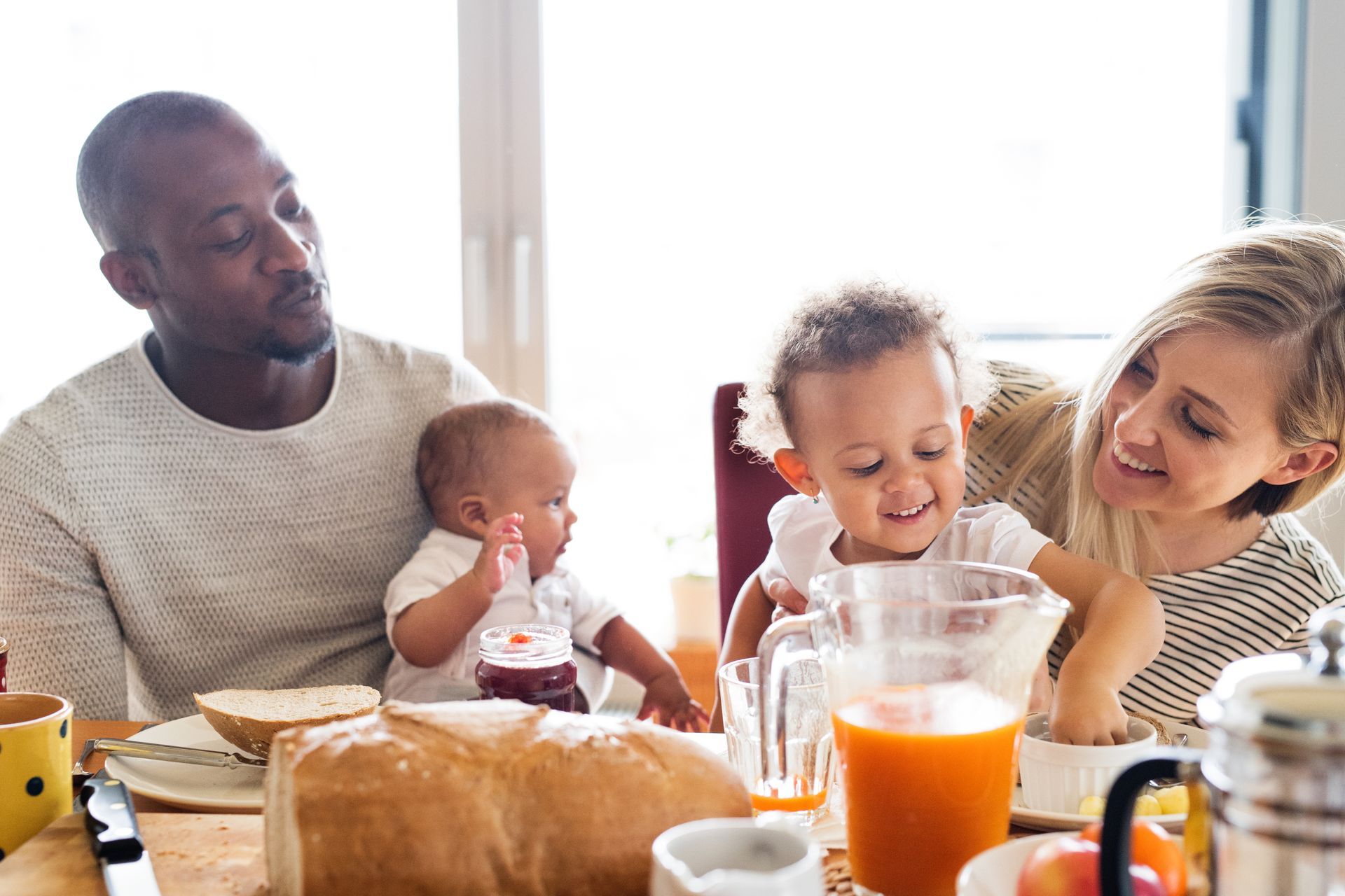 Family sharing breakfast at a table, with bread, juice, and smiling children in a bright kitchen
