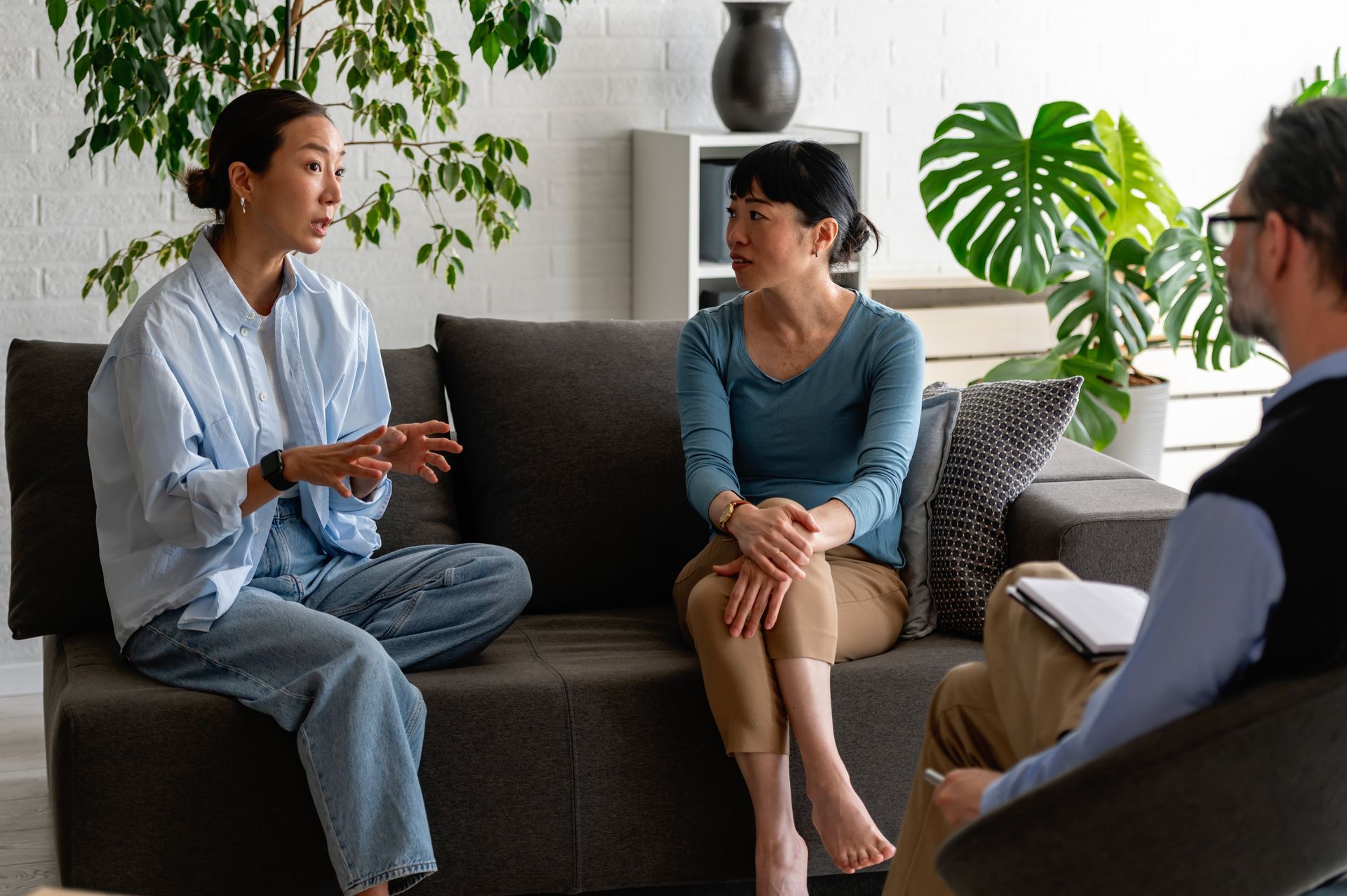 Three people talking in a bright living room, seated on a dark sofa with plants nearby.