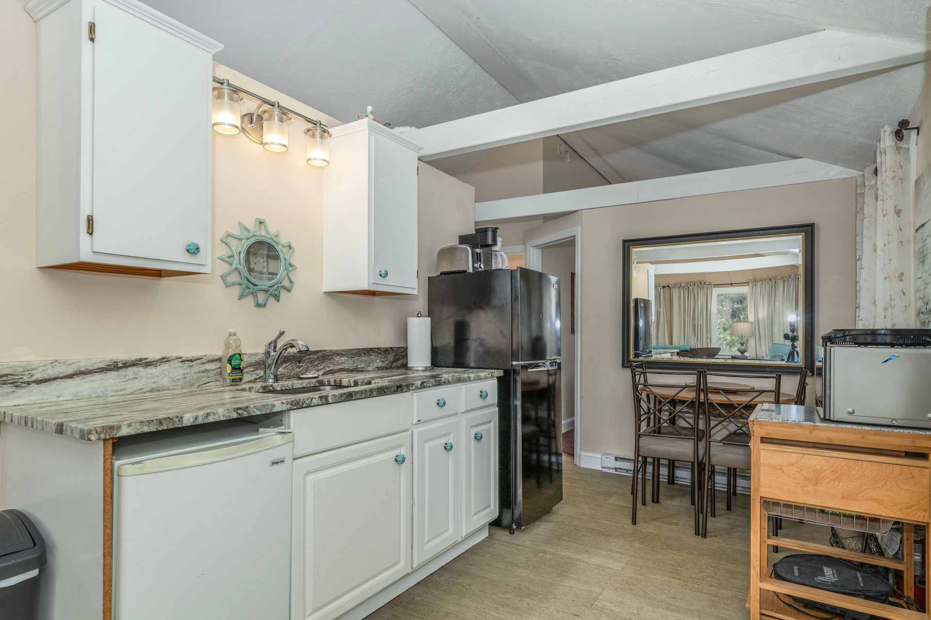 Small kitchen with white cabinets, black fridge, and dining area with mirror.