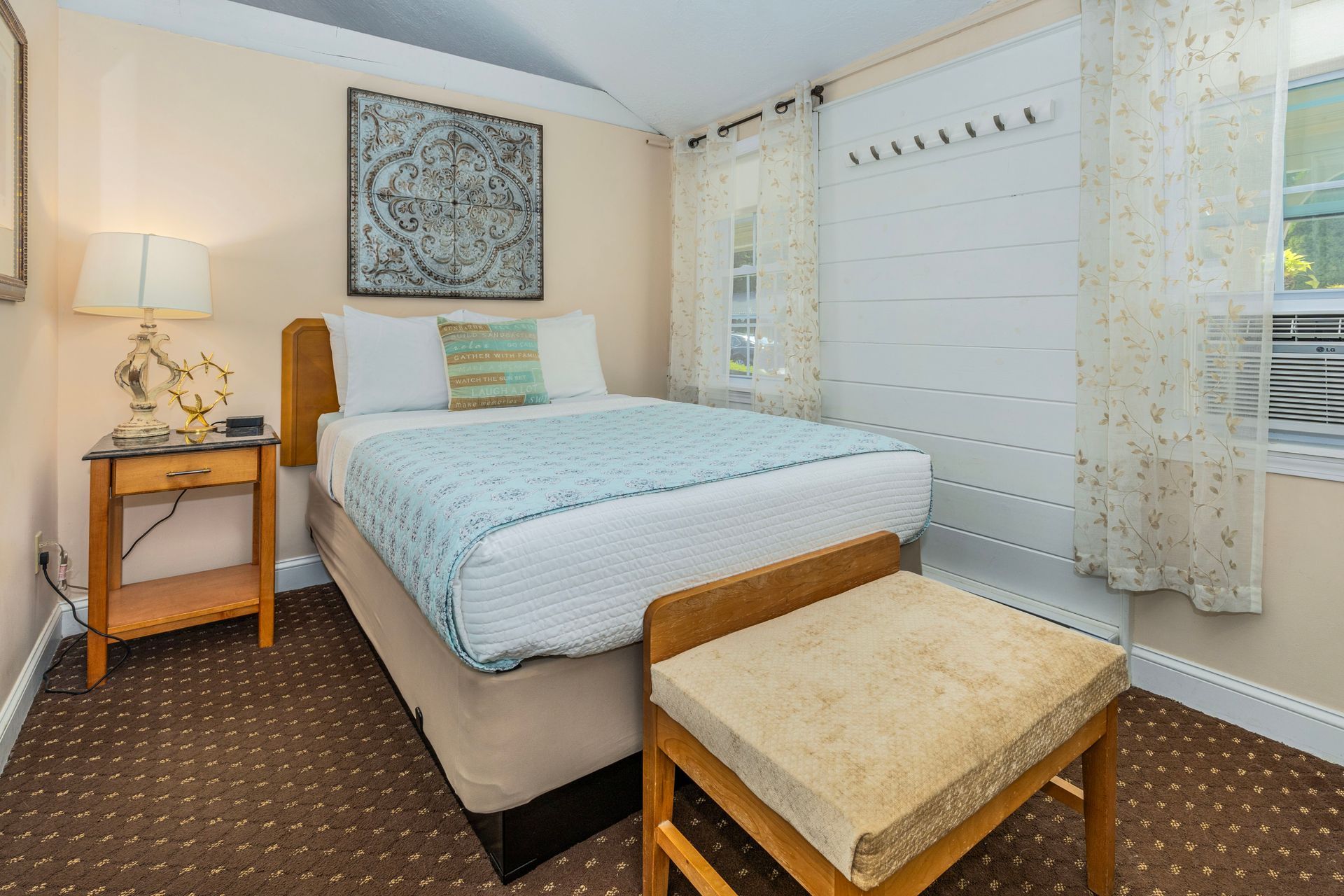 Cozy hotel room with a bed, nightstand, stool, and window with sheer curtains.