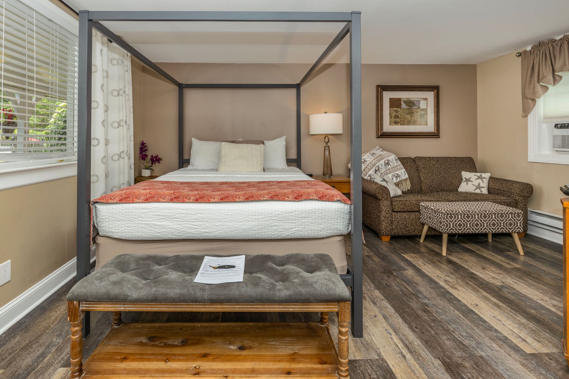 Bedroom with a four-poster bed, bench, sofa, and window. Beige walls, wood-look floor.