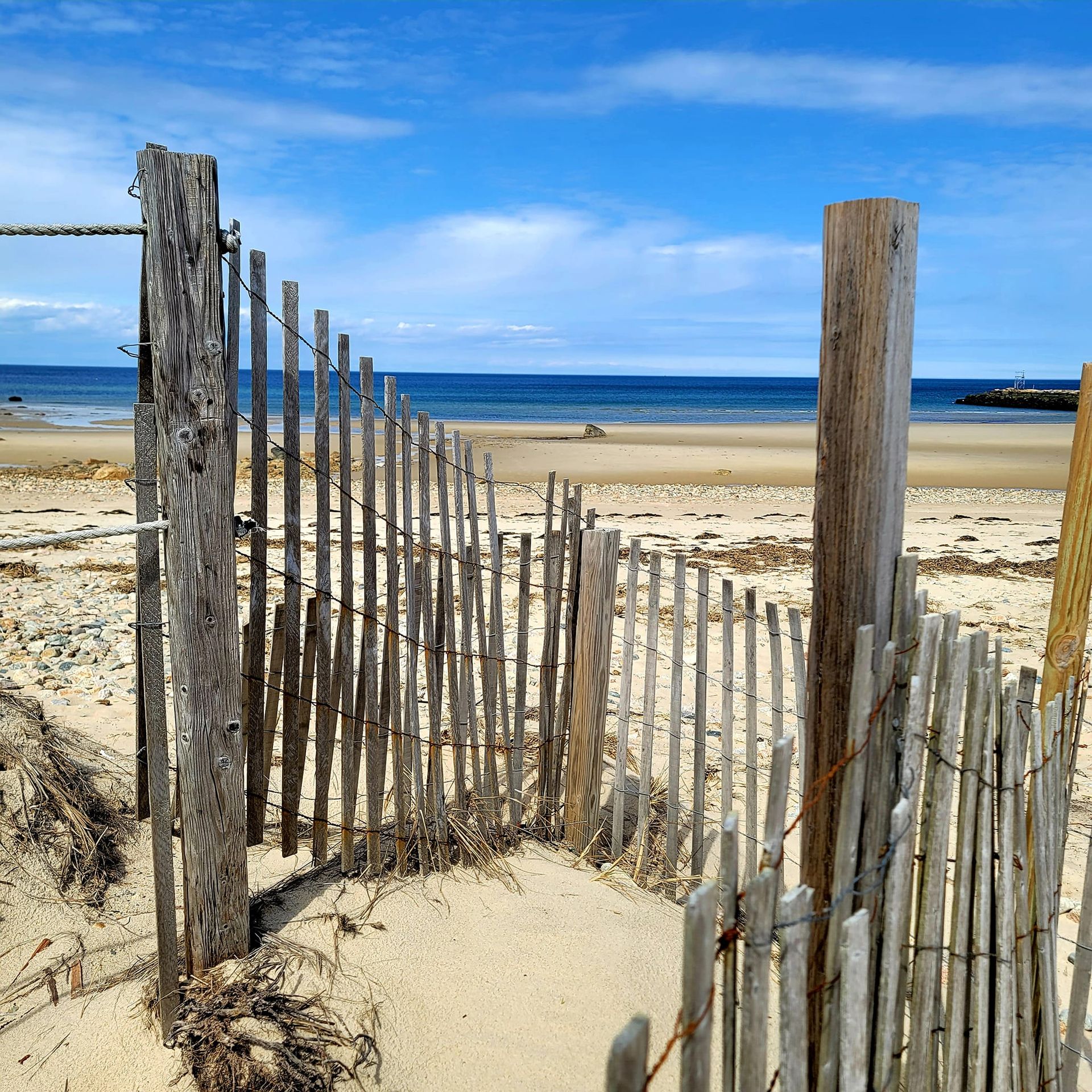 A weathered wooden fence on a sandy beach, with the ocean and blue sky in the background.