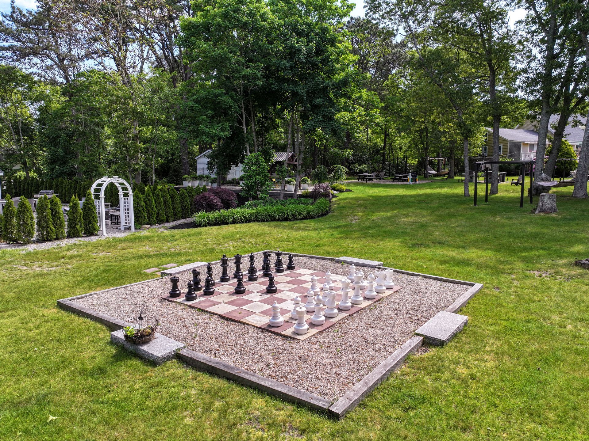 Outdoor chess set on a gravel square within a green lawn, surrounded by trees and landscaping.
