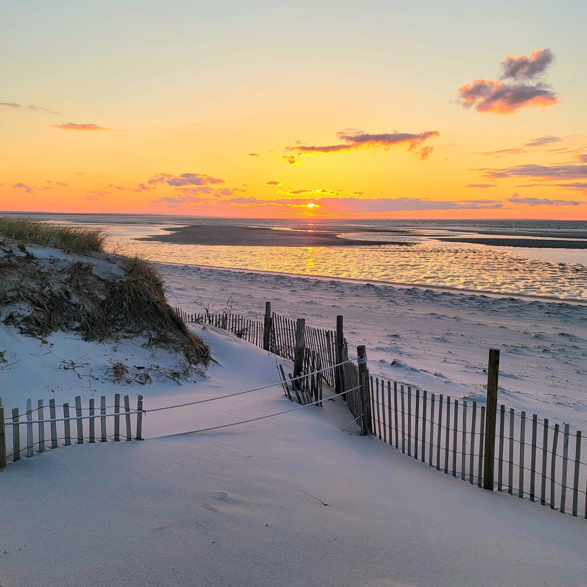 Sunset over a snowy beach, with a wooden fence and sand dune in the foreground.