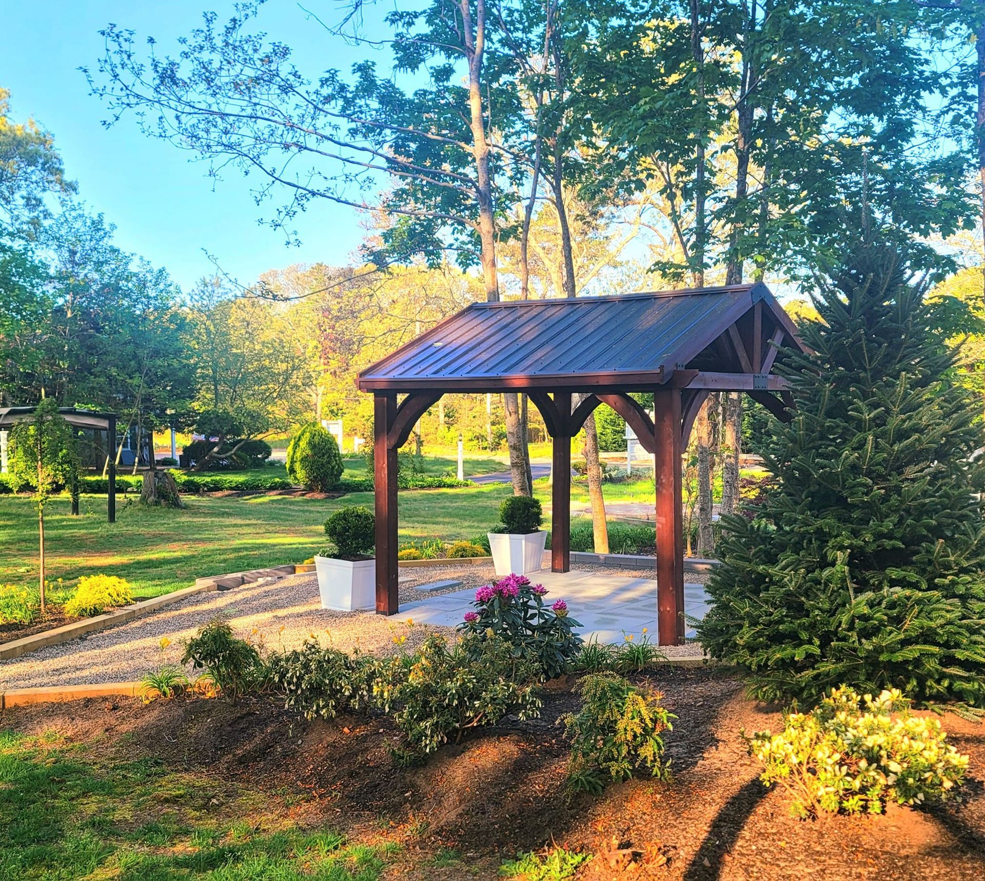 Wooden gazebo in a garden setting with trees, plants, and pathway. Two potted plants and greenery surround.