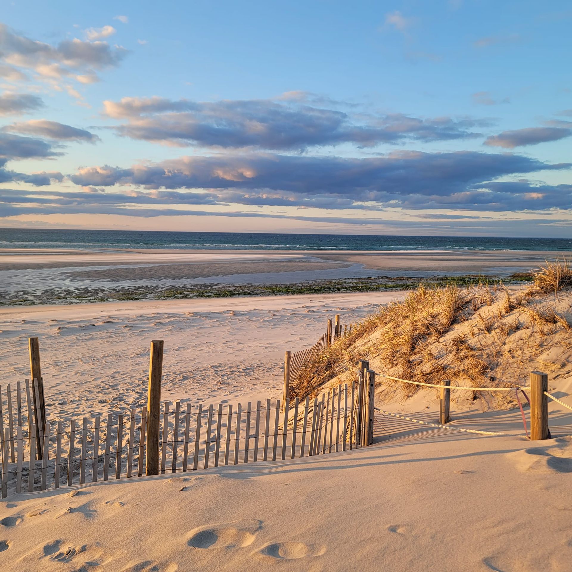 Sandy beach at sunset with a wooden fence and dunes. Calm ocean and cloudy sky in the background.