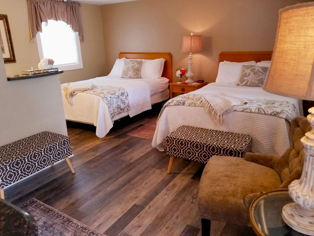 Bedroom with two twin beds, a patterned bench, and an armchair. Beige walls, wooden floors.