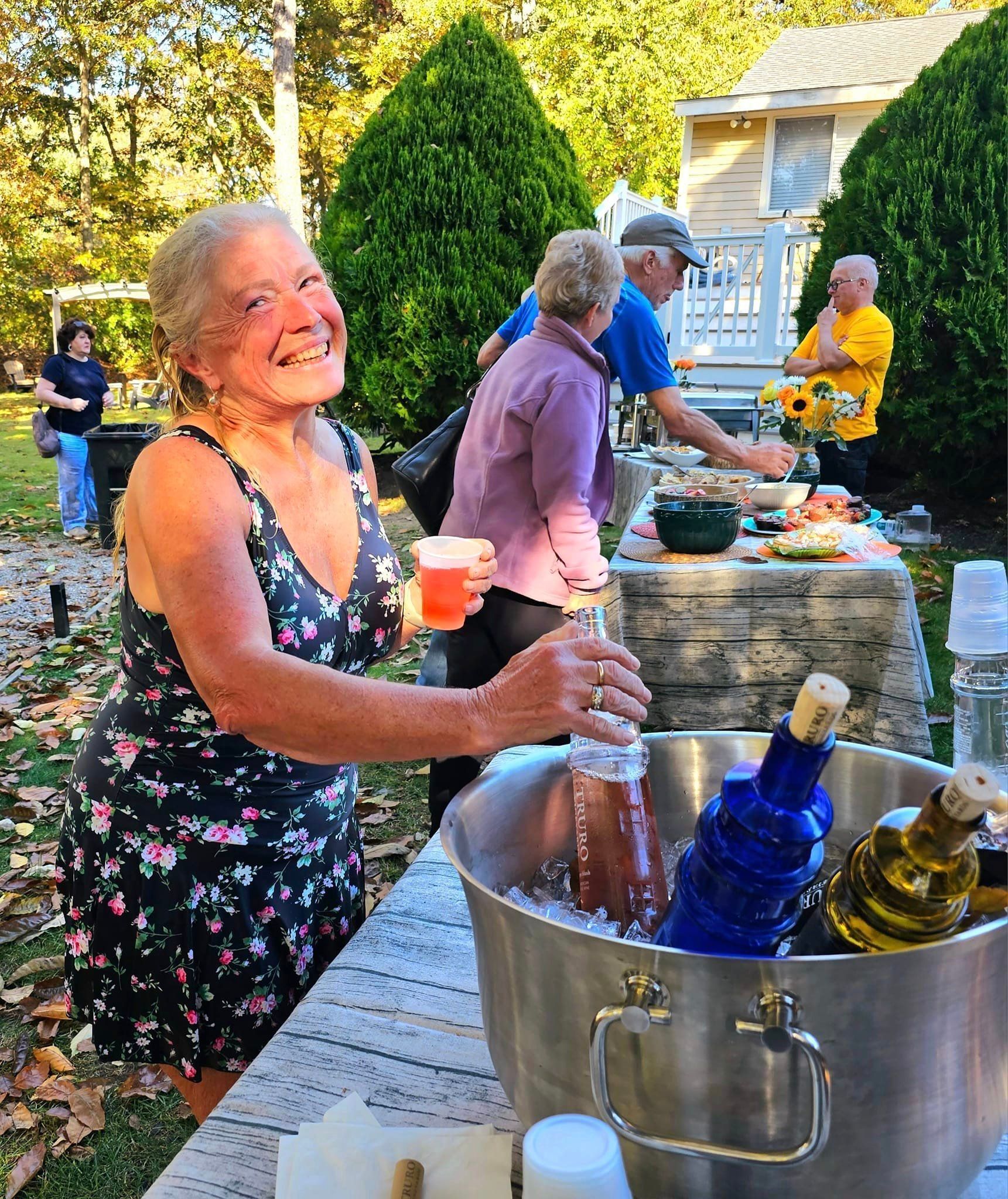Woman smiling at outdoor party, holding drink near ice bucket with bottles; others at food table.