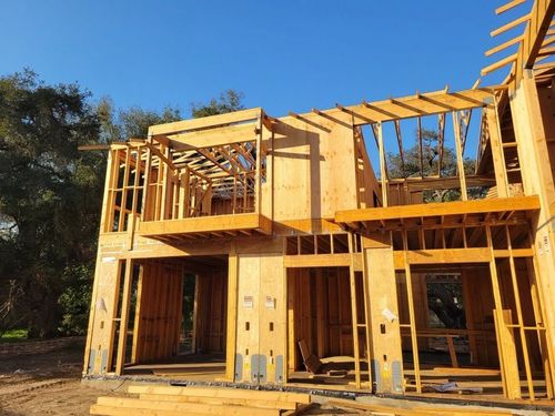 Two-story wooden house under construction with exposed framing and roof beams in bright sunlight