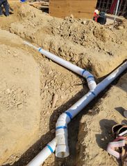 White PVC pipes with blue tape laid in a sandy trench at a construction site, with workers nearby