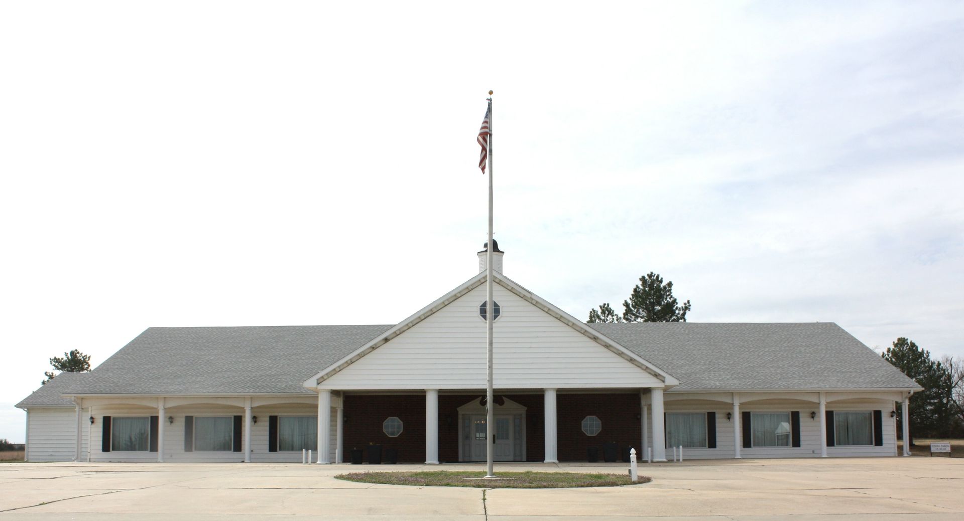 A wide, single-story building with a white gabled entrance, a large parking lot, and a tall flagpole in front.