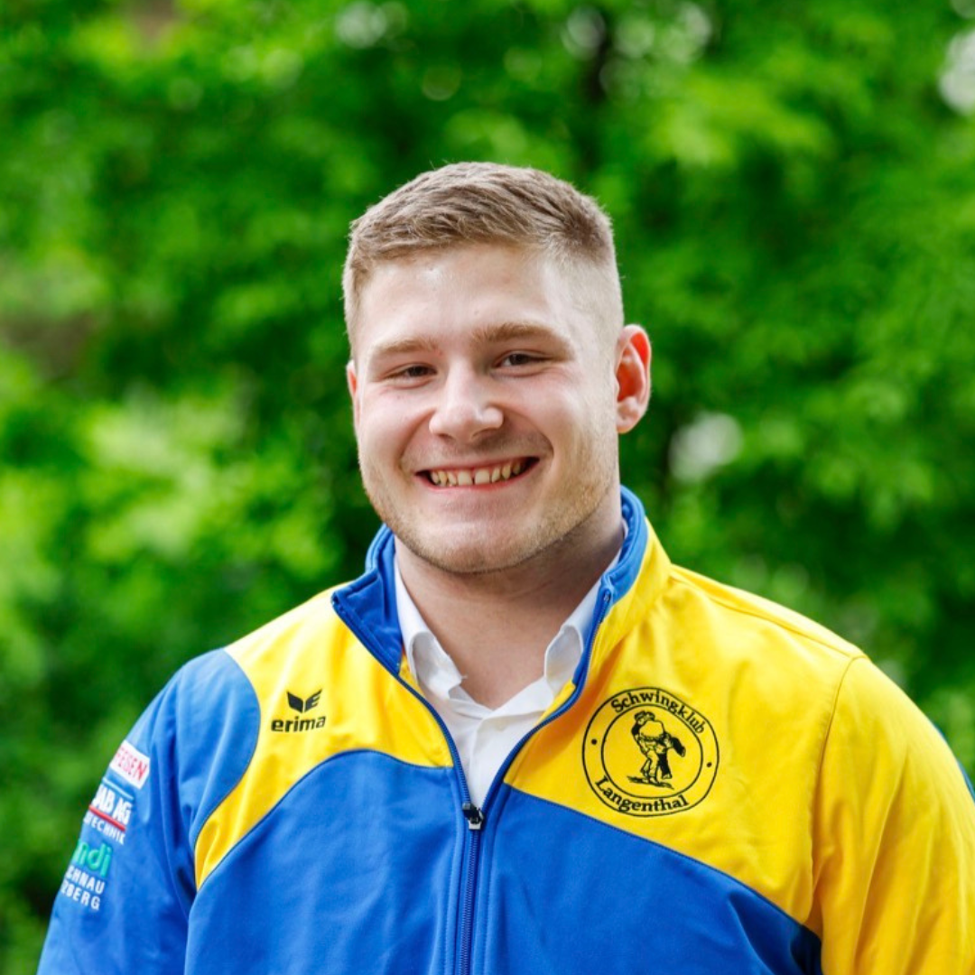 Ein Mann in gelb-blauer Jacke lächelt vor grünem Hintergrund.