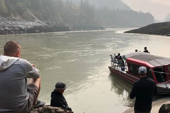 A group of people are sitting on the shore of a river looking at a boat.