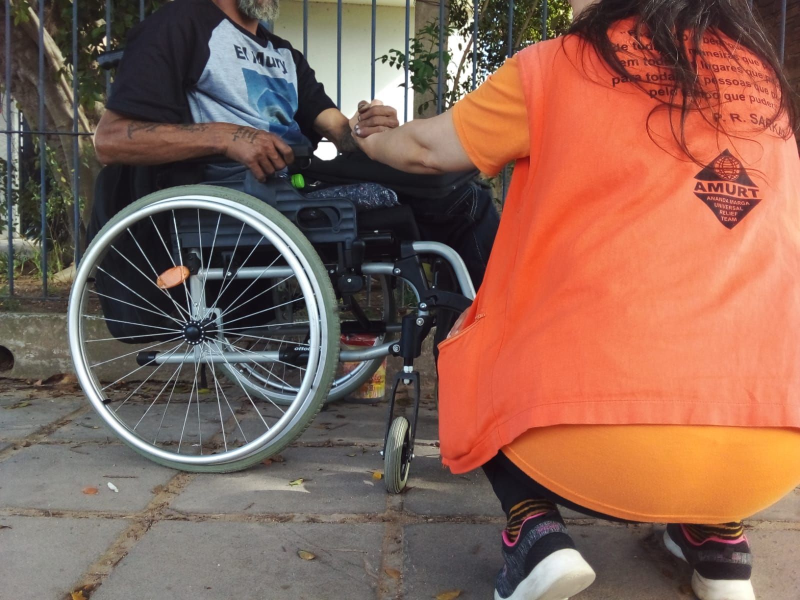 Pessoa em cadeira de rodas auxiliada por uma pessoa vestindo um colete laranja; eles estão do lado de fora.