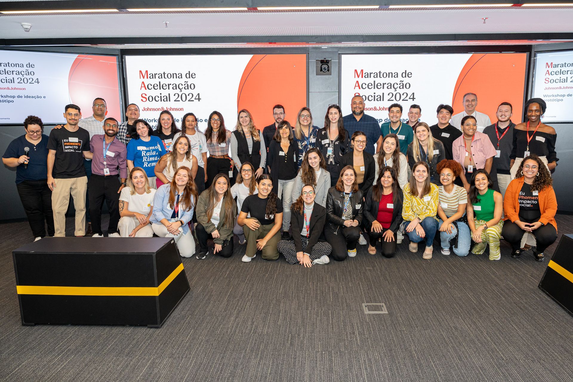 Um grupo de pessoas em pé numa sala de conferências em frente a telas que exibem a