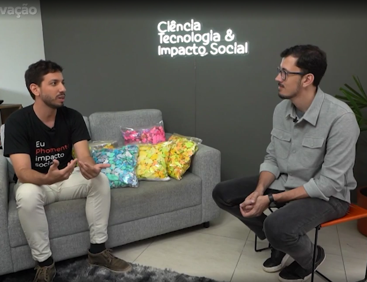 Dois homens sentados em um estúdio, conversando ao lado de um sofá cinza com almofadas coloridas e a inscrição “Ciência, tecnologia e impacto social”.