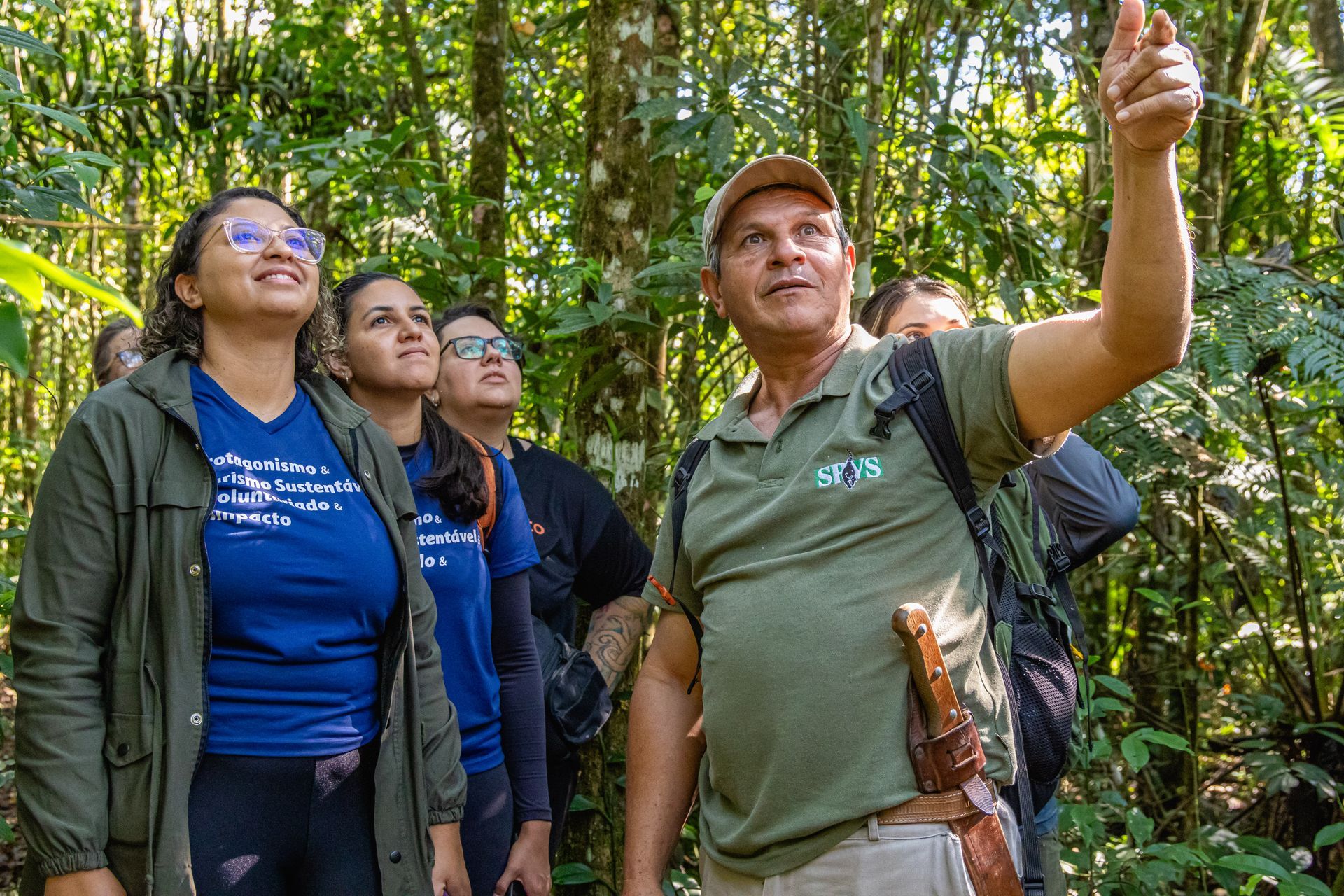 Um grupo de pessoas em uma floresta ouve um guia que aponta para as árvores, com a luz do sol filtrando-se através das folhas.