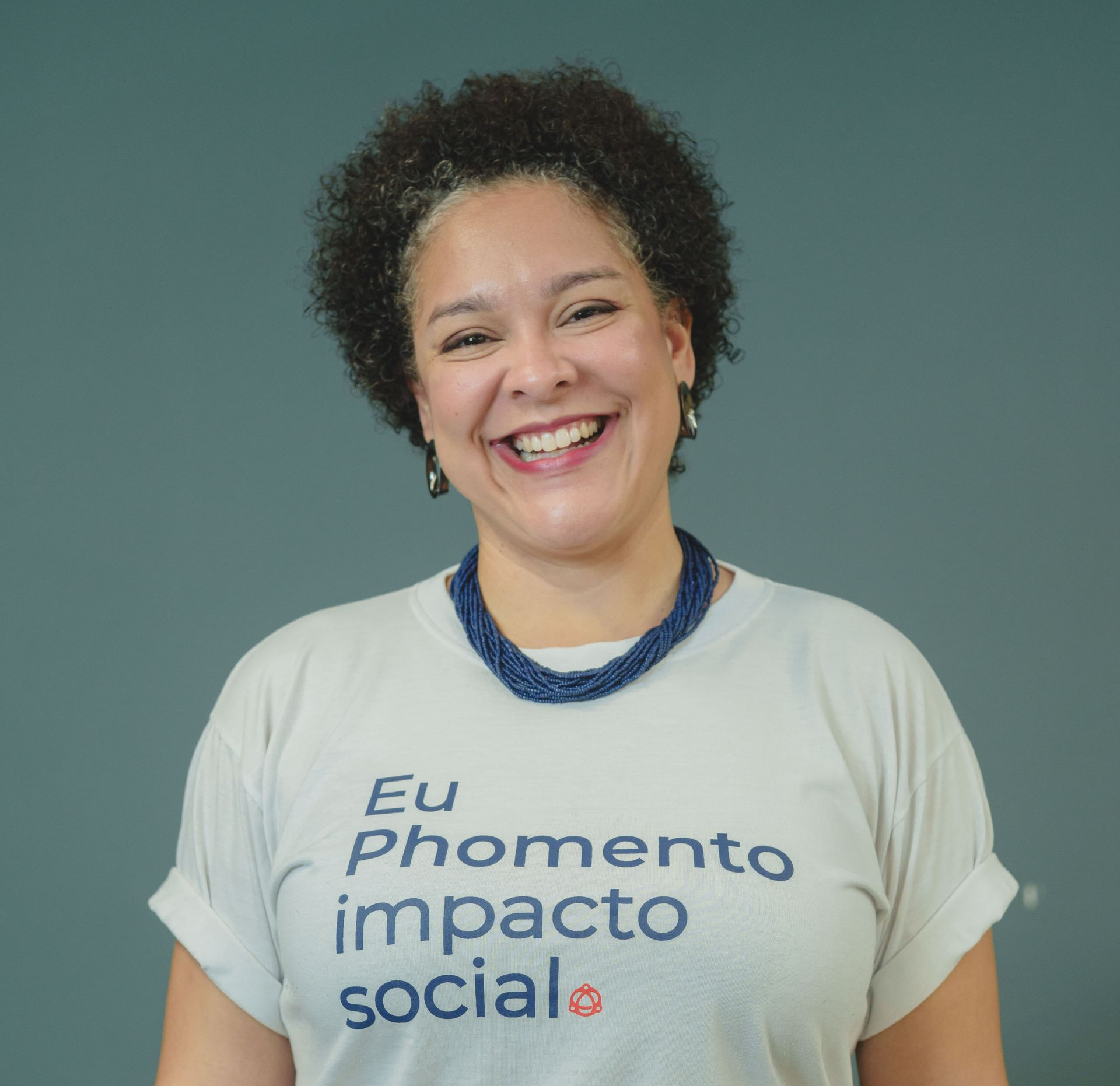Pessoa sorridente vestindo uma camiseta branca com a inscrição “Eu Phomento impacto social” e um colar azul contra um fundo verde.