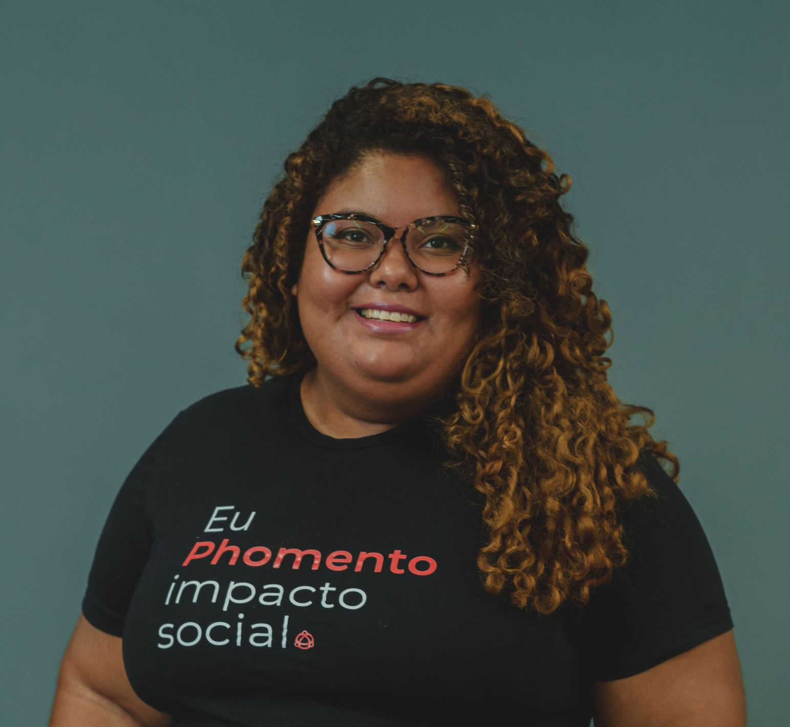 Mulher sorridente de óculos e camiseta preta com a frase “Eu fomento impacto social” sobre um fundo verde-azulado.