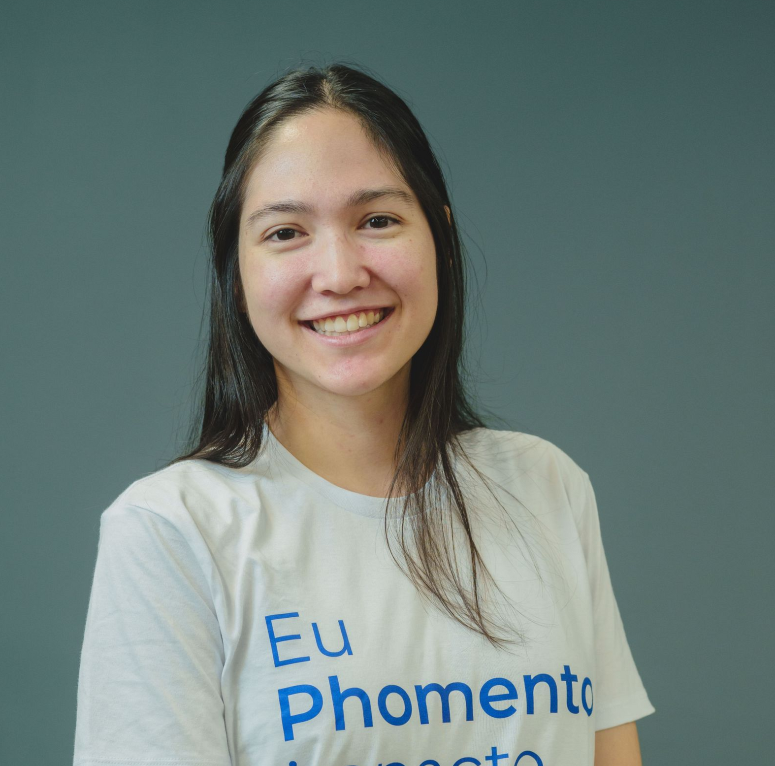 Pessoa sorridente vestindo uma camiseta branca com estampa azul, em pé contra um fundo cinza liso.