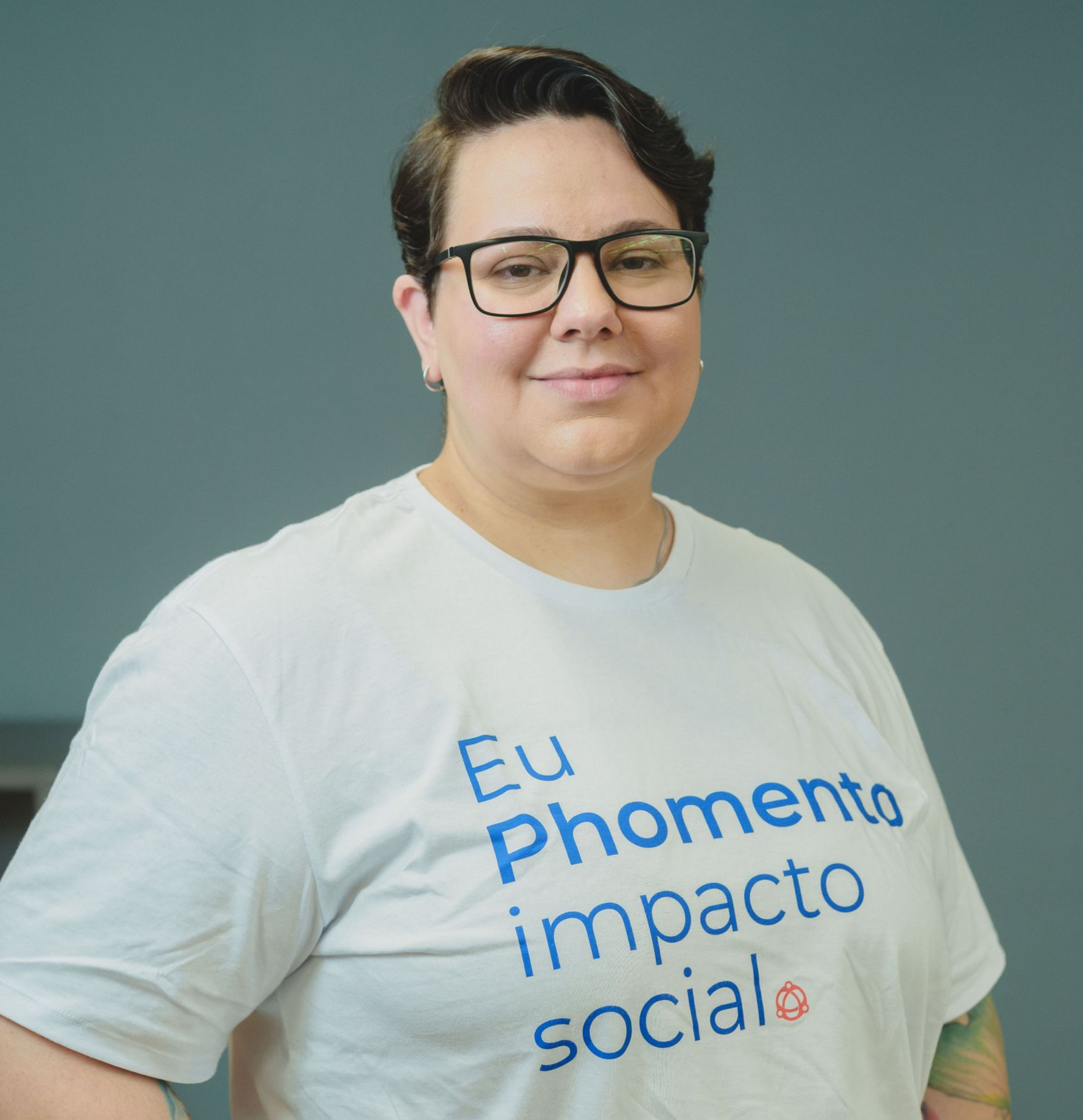 Pessoa de óculos vestindo uma camiseta branca com estampa azul, em pé contra um fundo cinza.