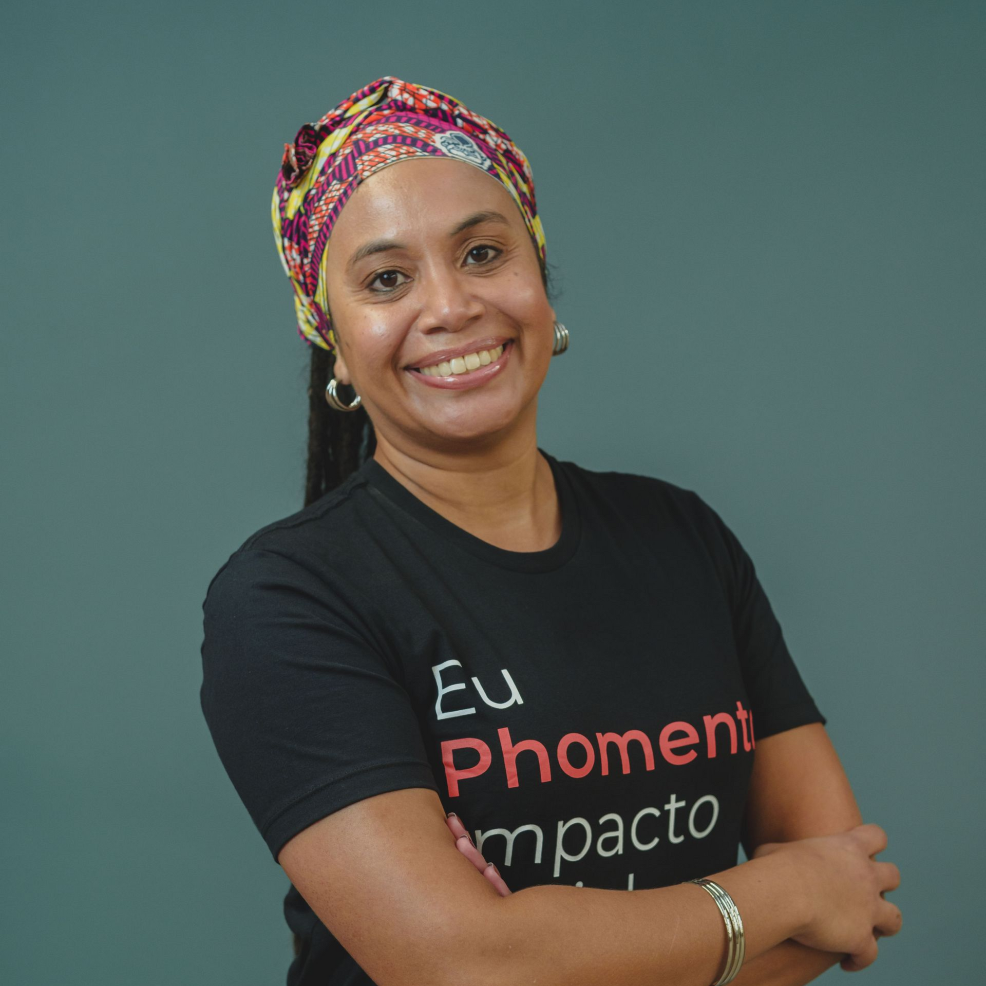 Mulher sorridente com um turbante colorido e camiseta preta, em pé com os braços cruzados contra um fundo azul-petróleo.