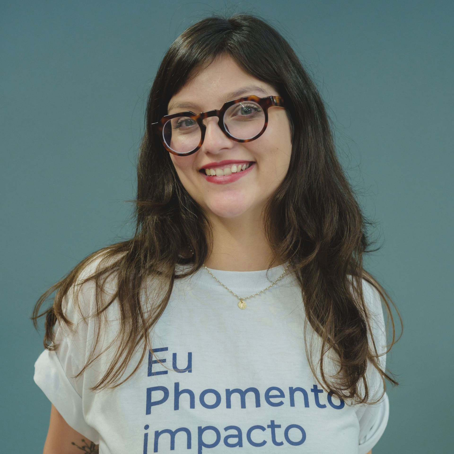 Mulher de óculos e camiseta branca com a inscrição “Eu Phomento Impacto”, sorrindo contra um fundo azul-petróleo.