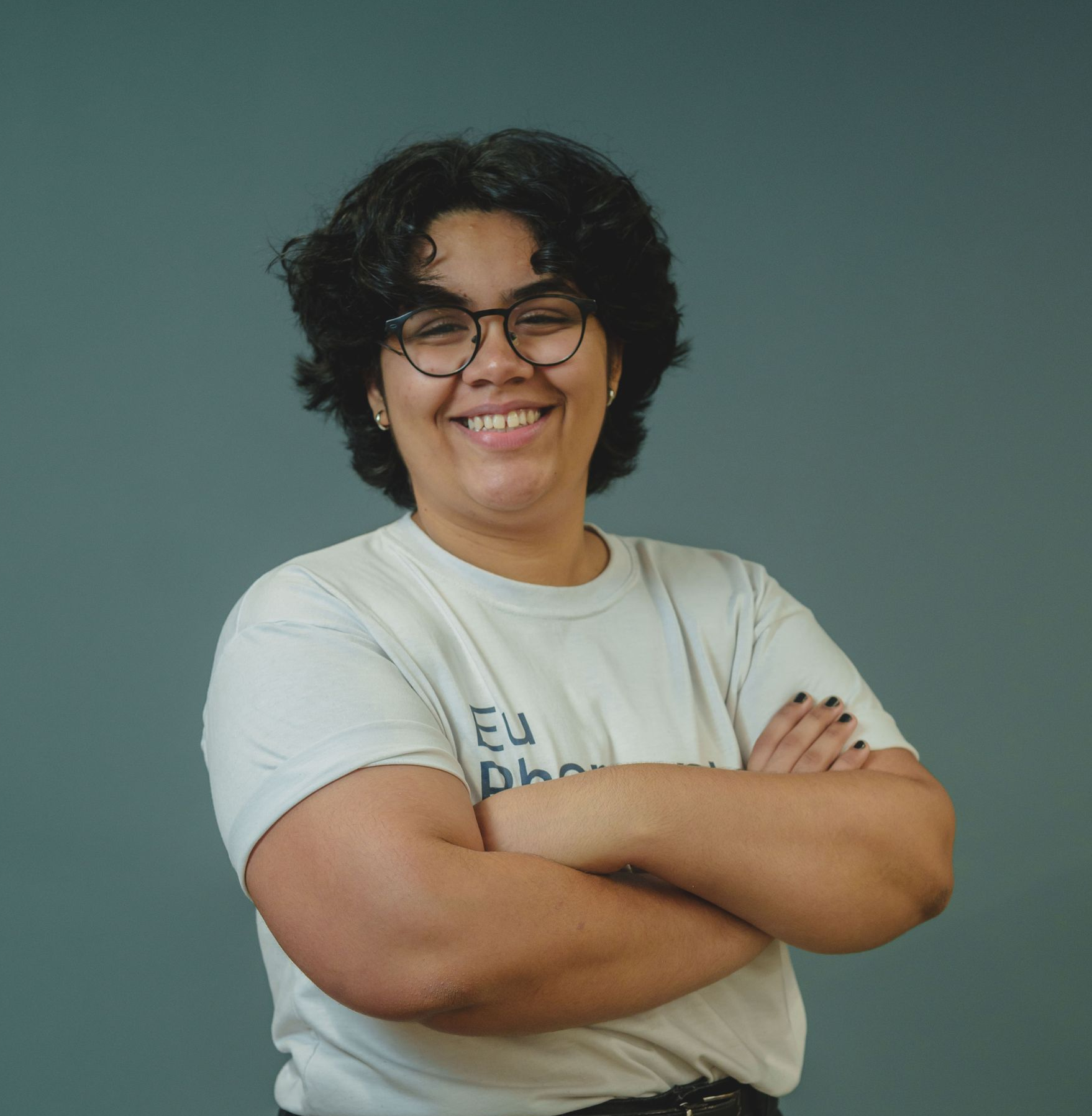 Pessoa sorridente com cabelo encaracolado e óculos, braços cruzados, vestindo uma camiseta branca sobre um fundo verde-azulado liso.
