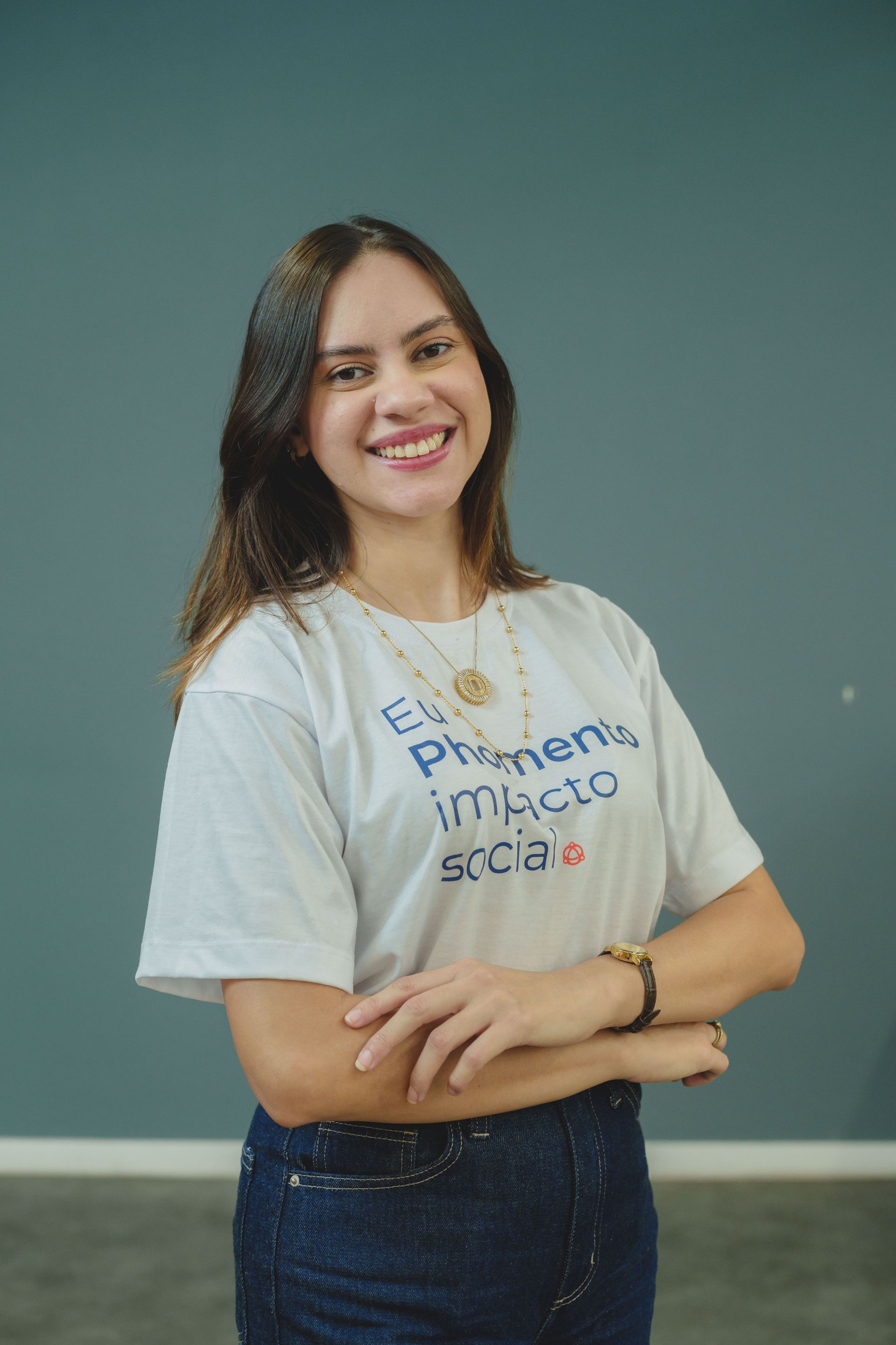 Mulher sorrindo com os braços cruzados, vestindo camiseta branca e calça jeans, contra uma parede verde-azulada.