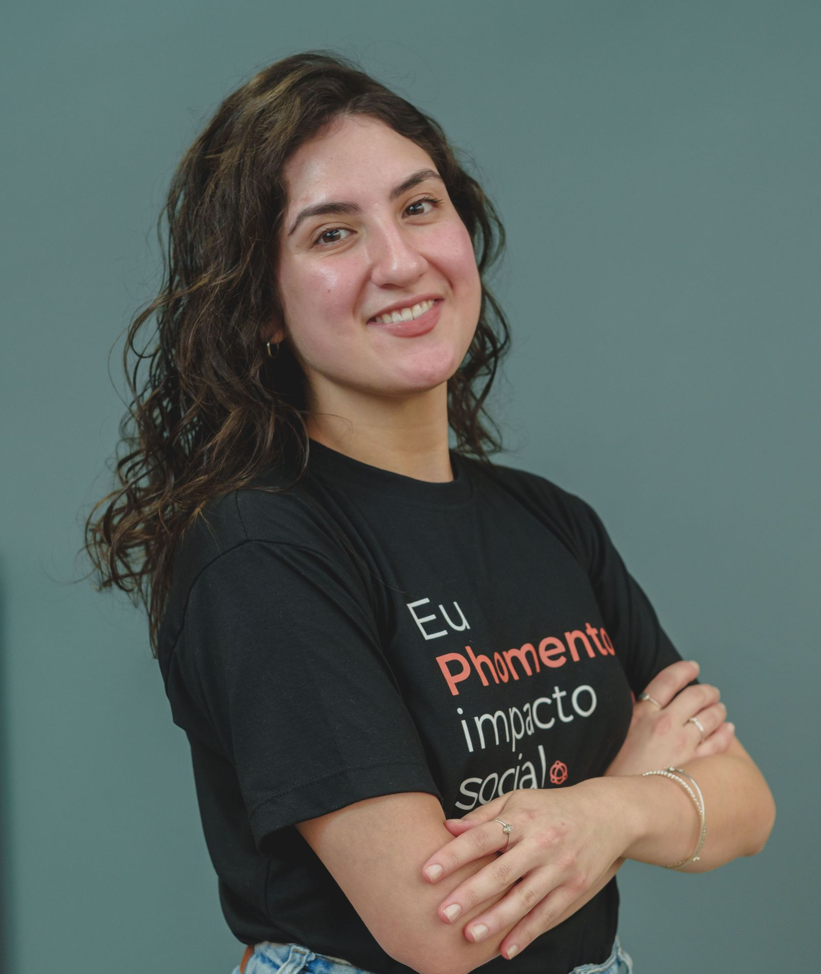 Pessoa sorridente vestindo camiseta preta, com os braços cruzados, em pé contra um fundo verde-azulado.