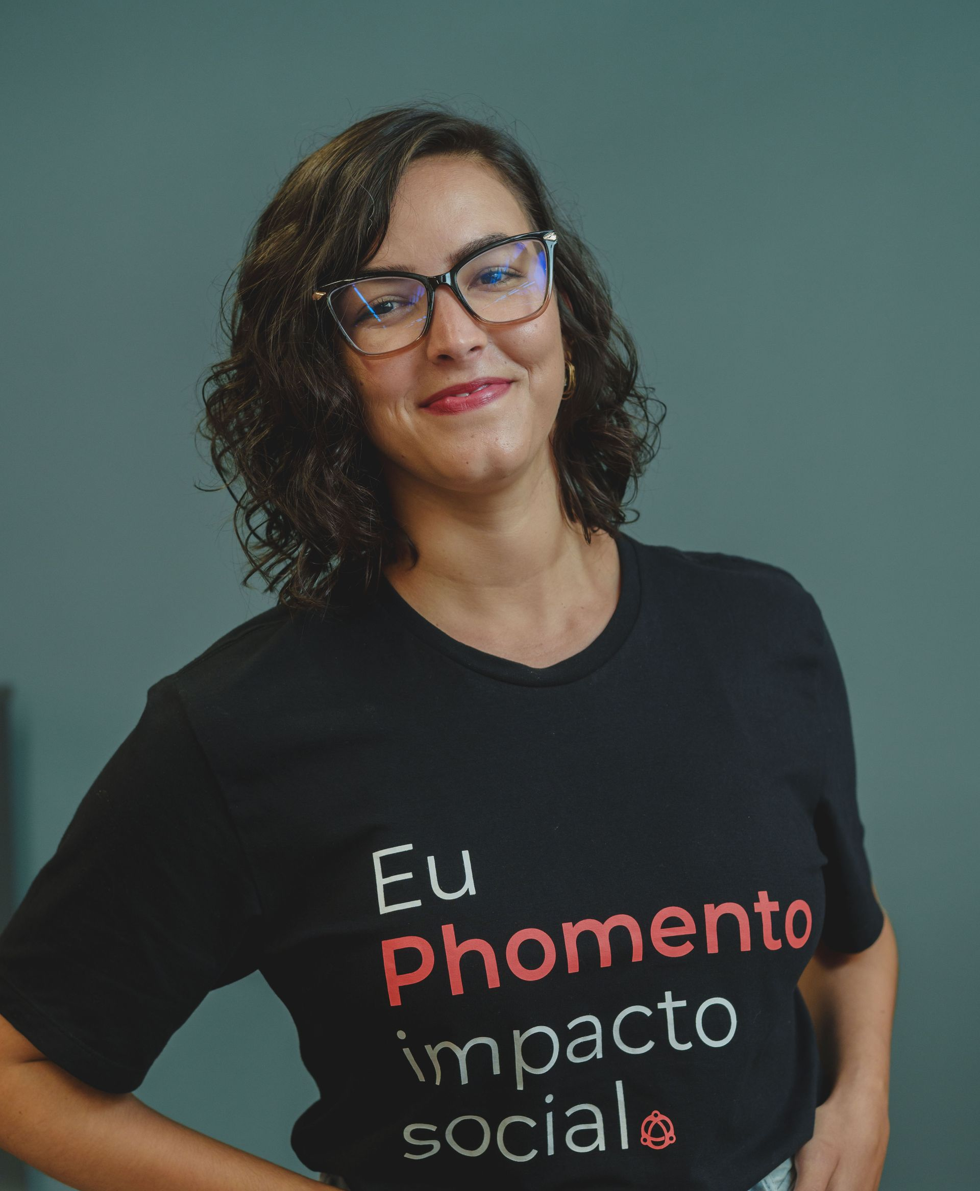 Mulher sorridente de óculos e camiseta preta com a inscrição “Eu phomento impacto social”, contra um fundo verde-azulado.