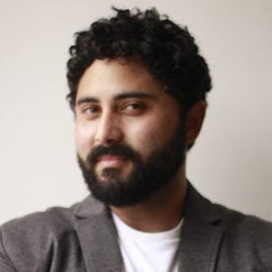Retrato de uma pessoa barbuda com cabelo escuro encaracolado, vestindo um blazer cinza e camisa branca, contra um fundo claro.