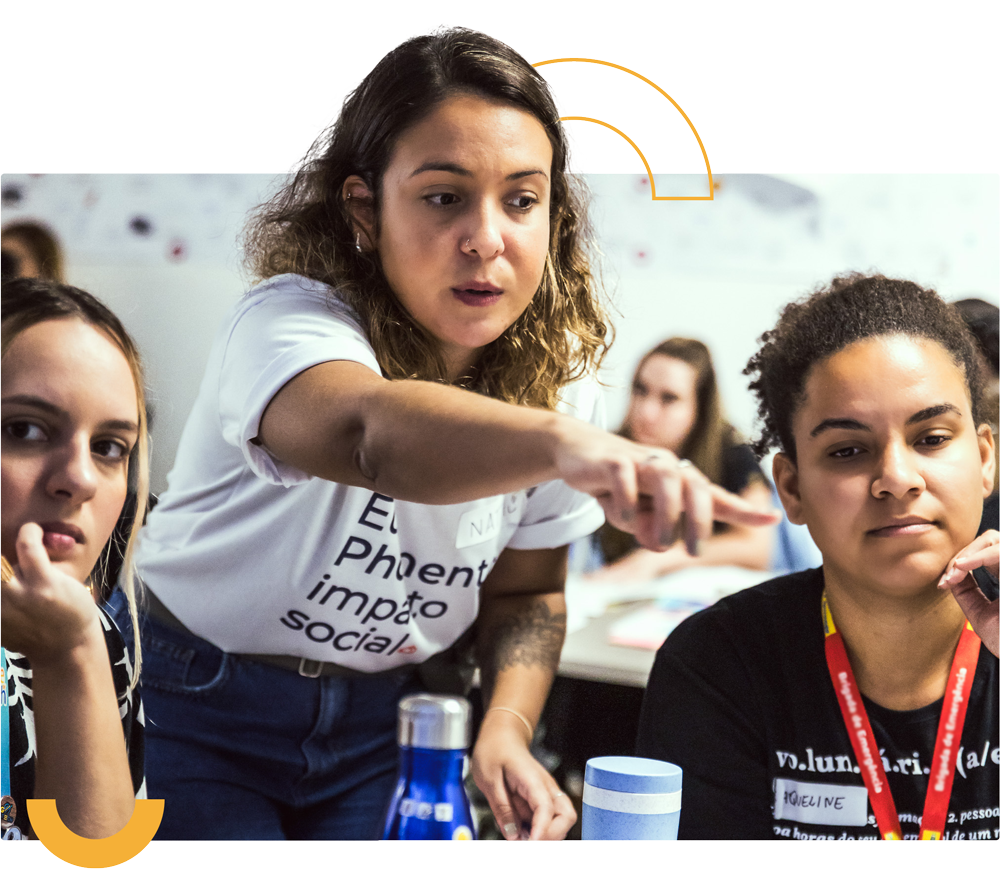 Uma mulher aponta para uma tela enquanto está de pé sobre um grupo de pessoas sentadas em carteiras em uma sala de aula.