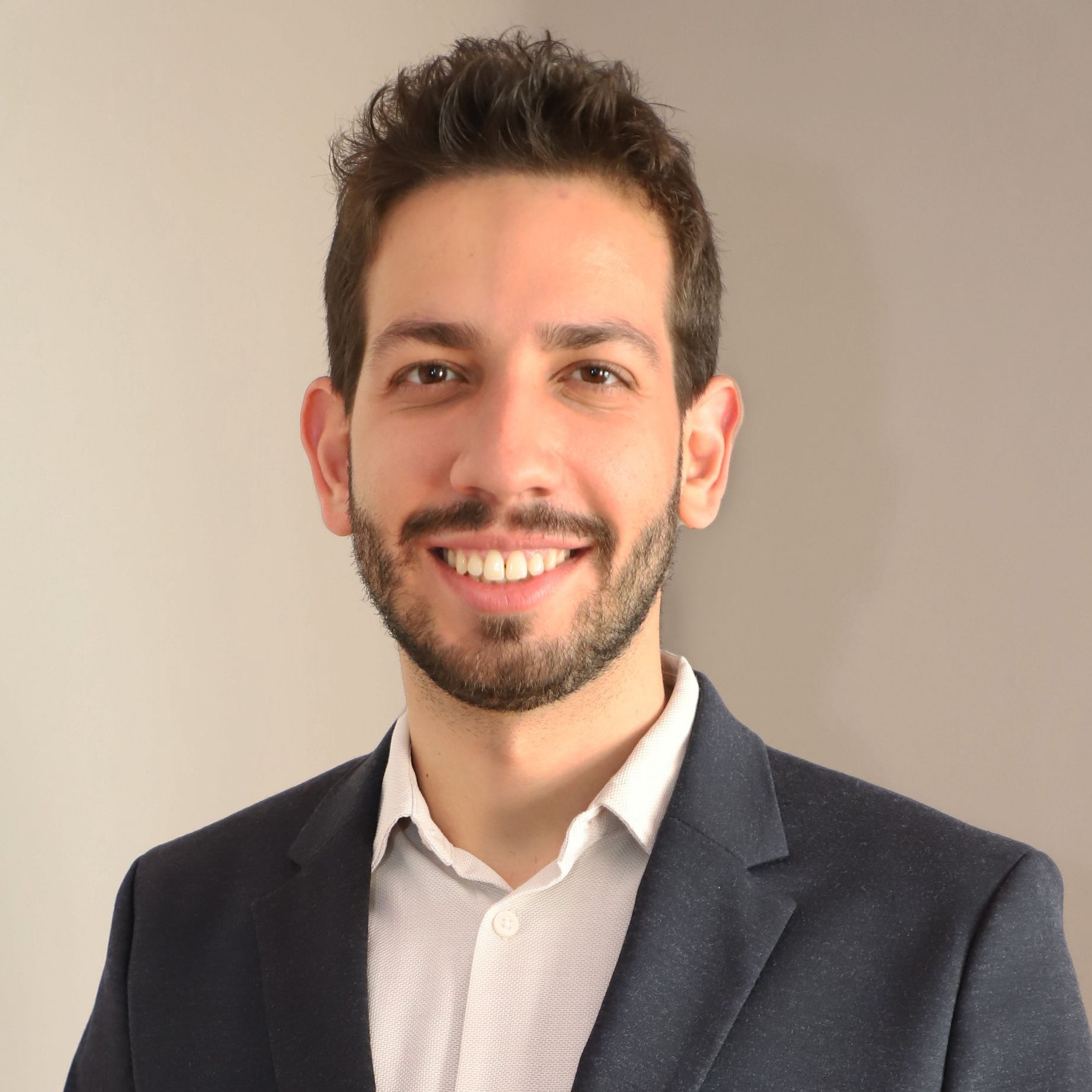 Homem sorridente de blazer escuro e camisa branca contra um fundo bege liso.