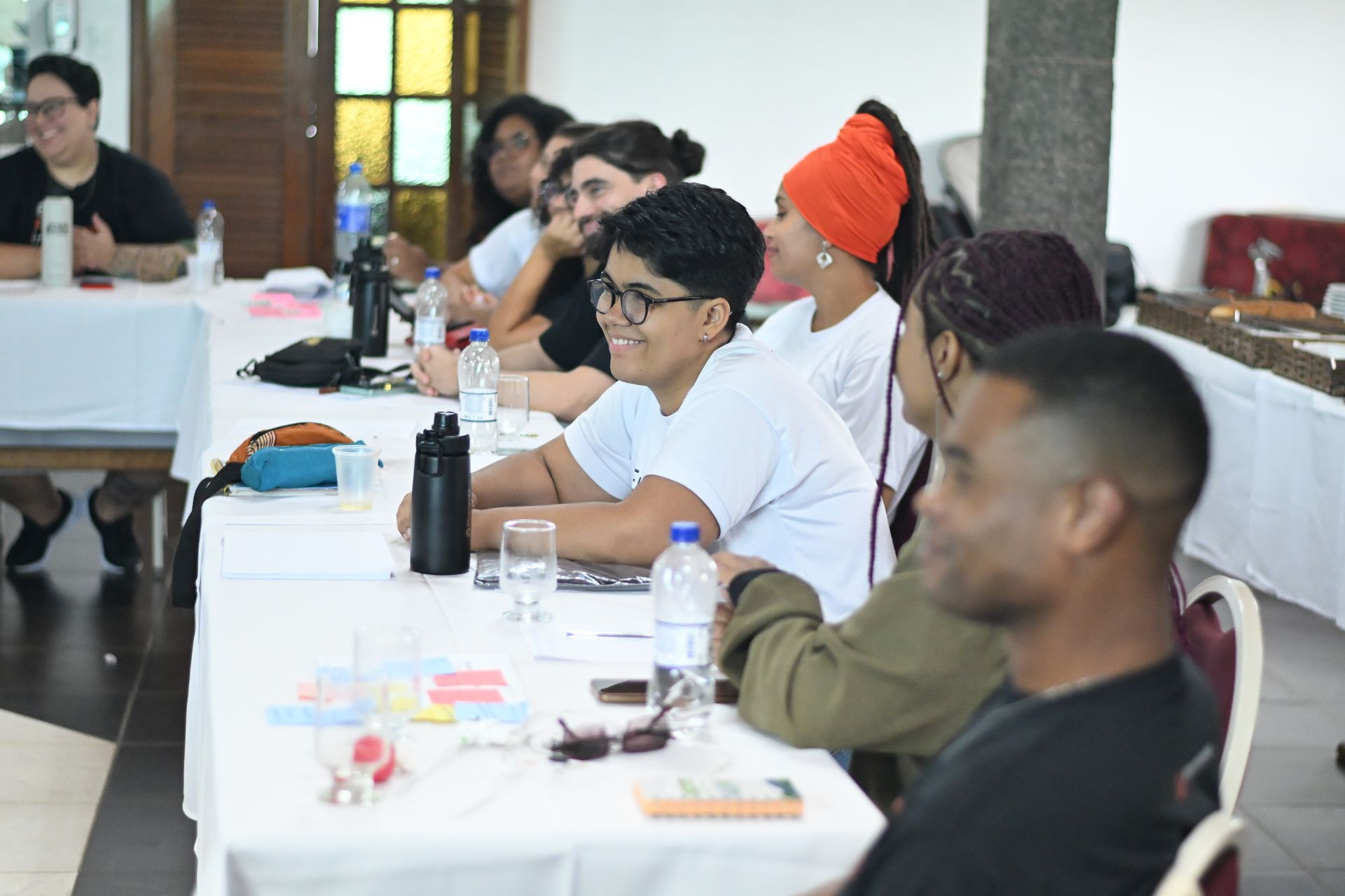 Um grupo de pessoas está sentado ao redor de uma longa mesa coberta com uma toalha branca, em uma sala bem iluminada, participando de uma discussão ou sessão de workshop.