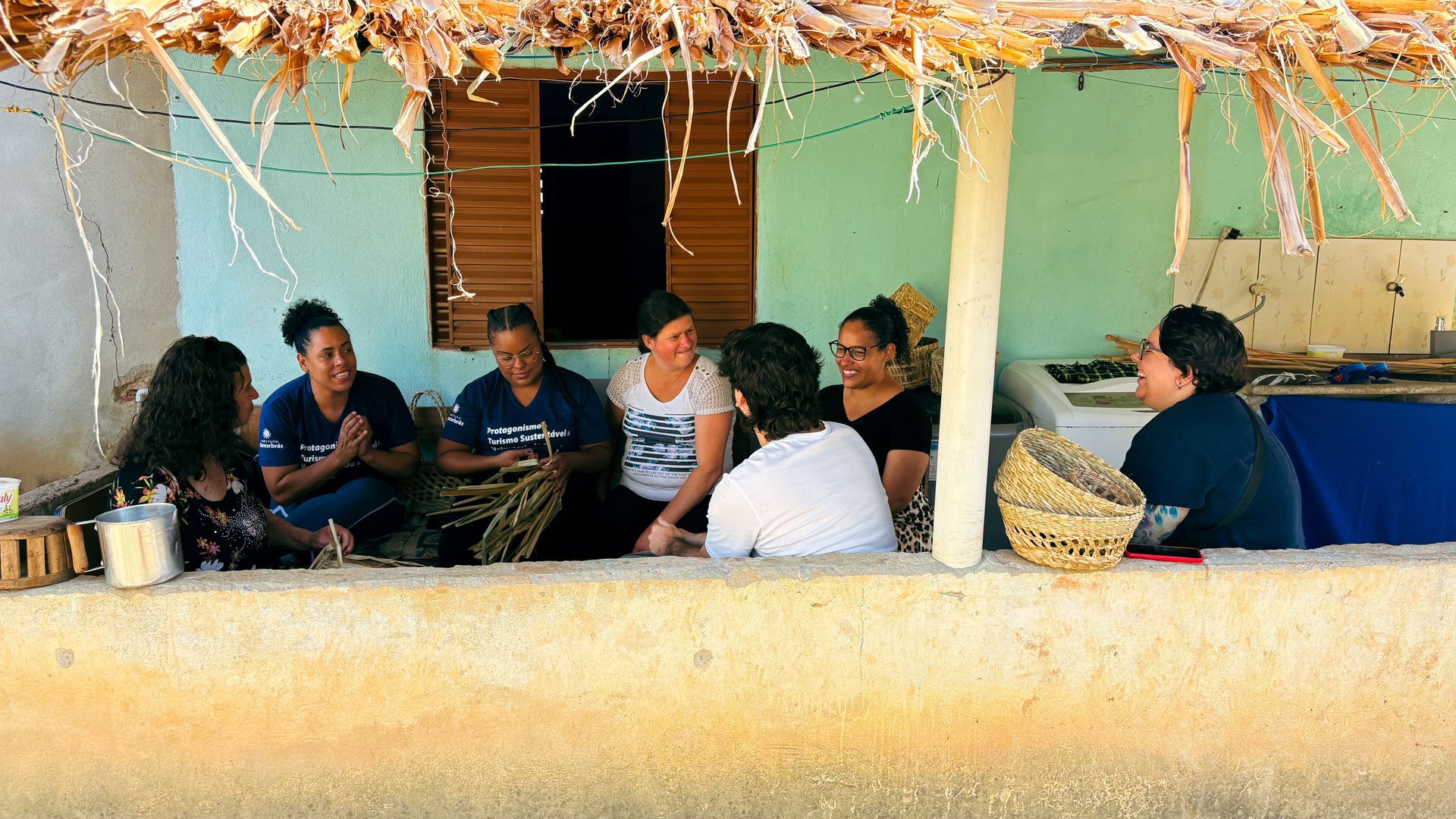 Um grupo de pessoas está sentado em círculo em uma varanda elevada com teto de palha, conversando.
