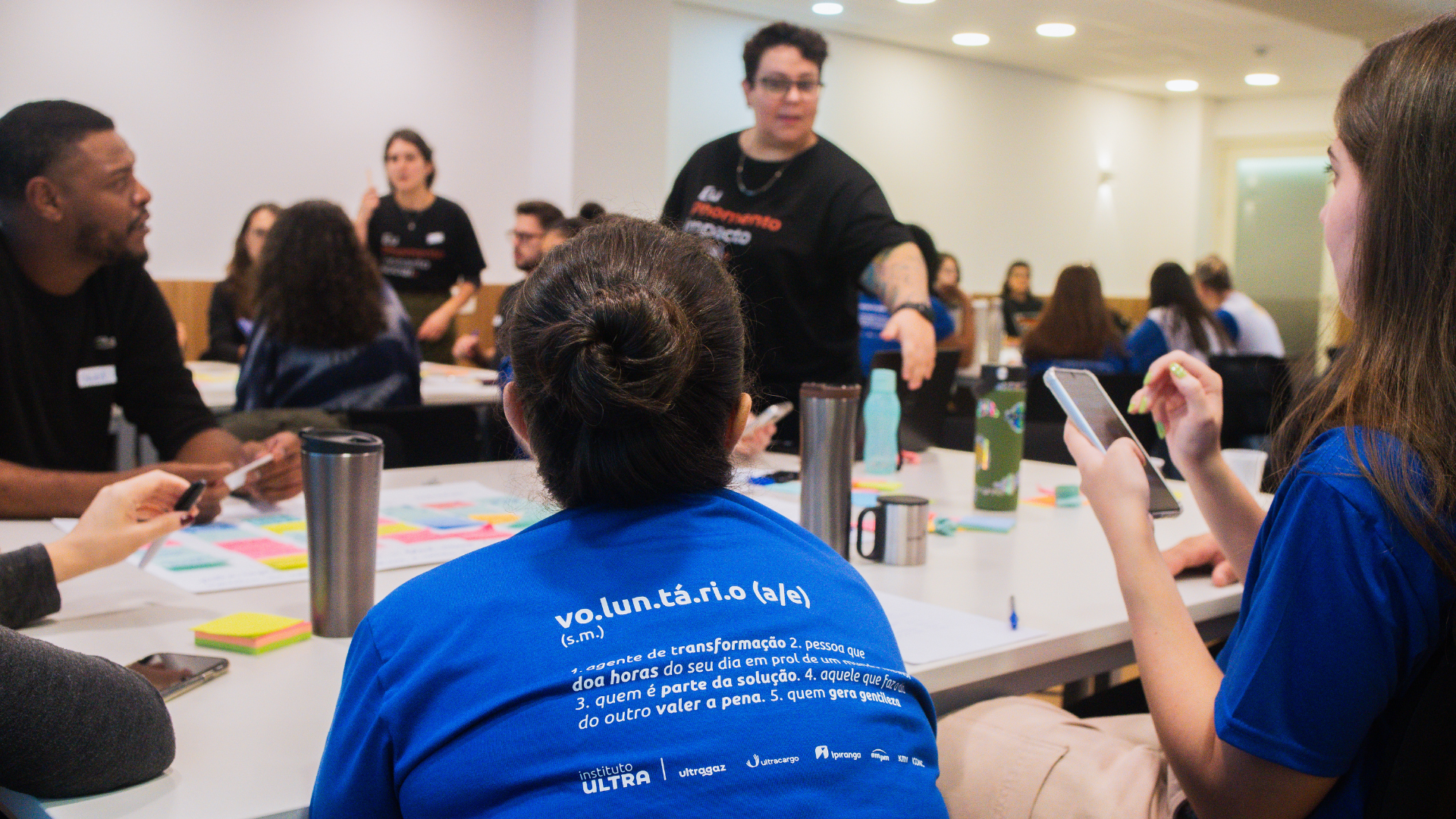 Um grupo de pessoas sentadas à mesa durante uma reunião ou workshop, com uma pessoa em pé gesticulando enquanto fala.