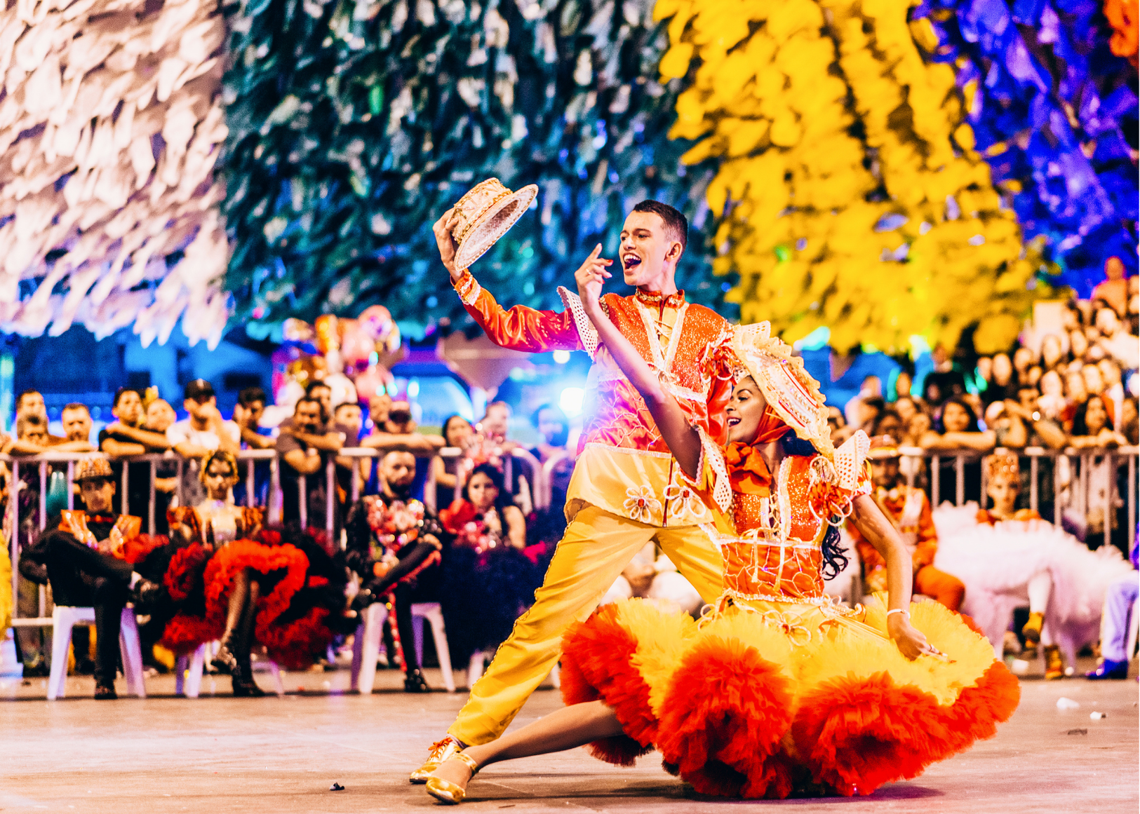 Uma dançarina com um traje amarelo e laranja vibrante se apresenta em um palco iluminado por luzes coloridas, diante de uma plateia sentada.