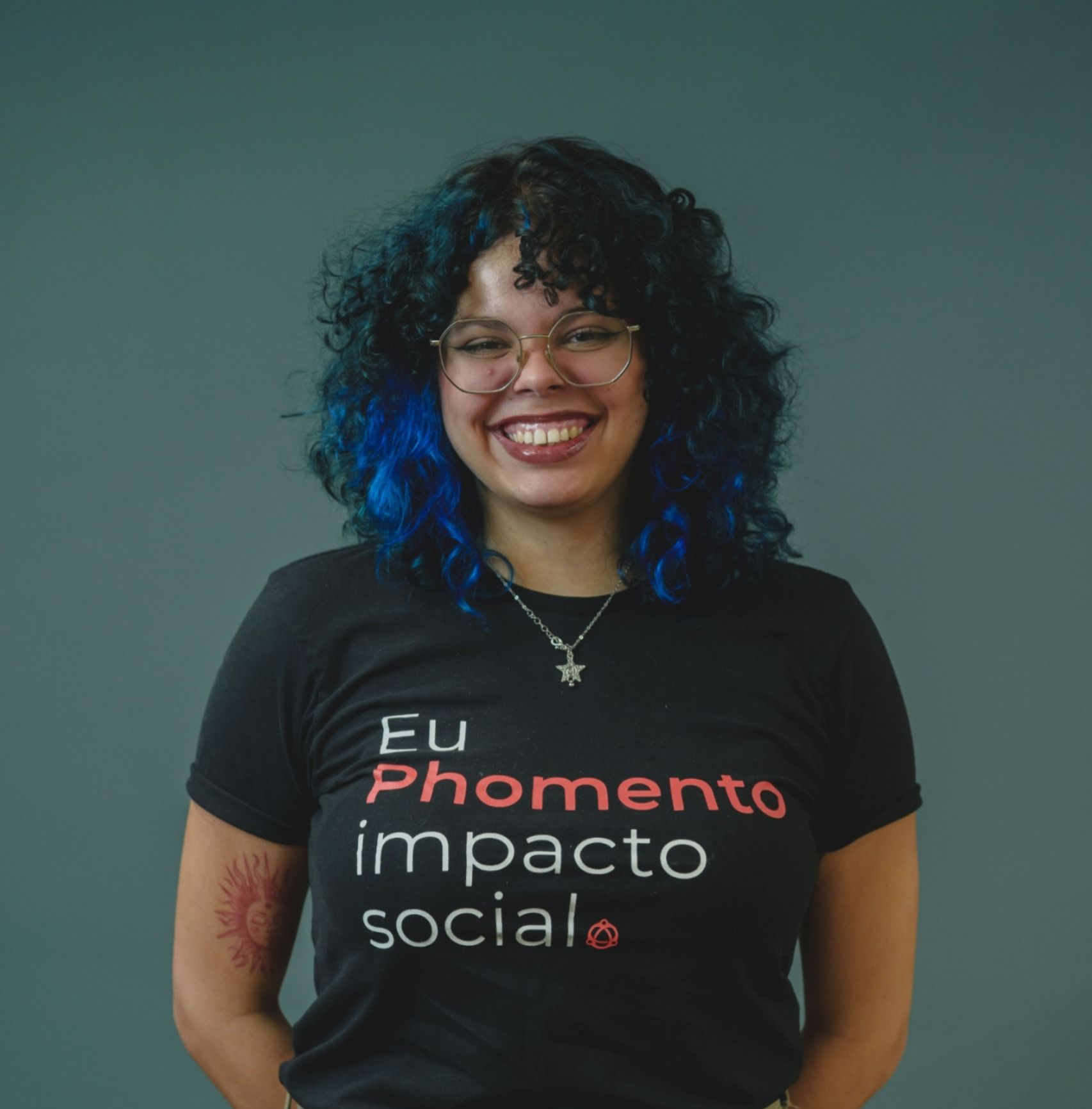 Pessoa sorridente com cabelo encaracolado vestindo uma camisa preta que diz “Eu Prometo impacto social”