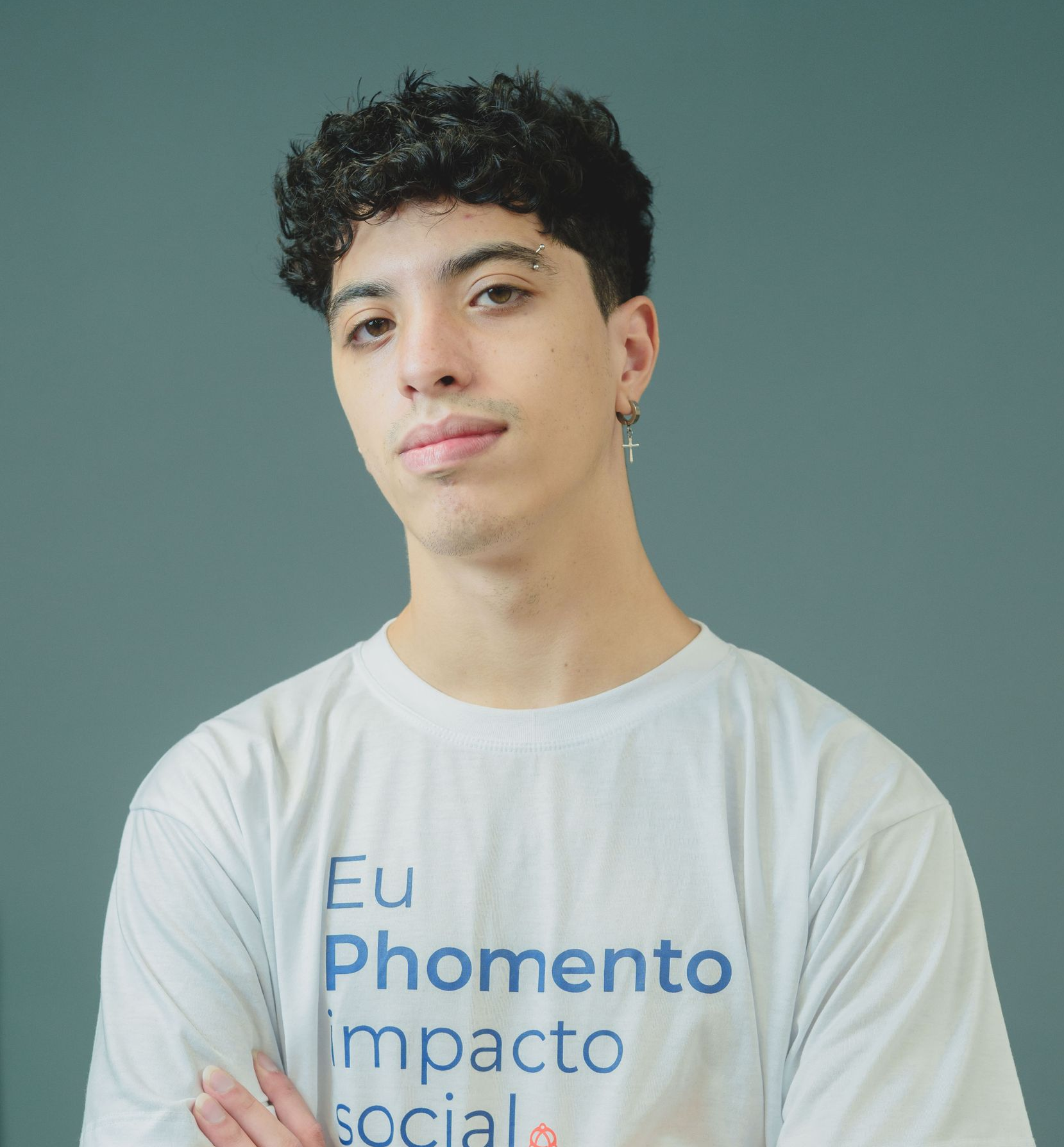 Jovem vestindo camiseta branca com estampa azul, em pé contra um fundo verde-azulado com expressão neutra.