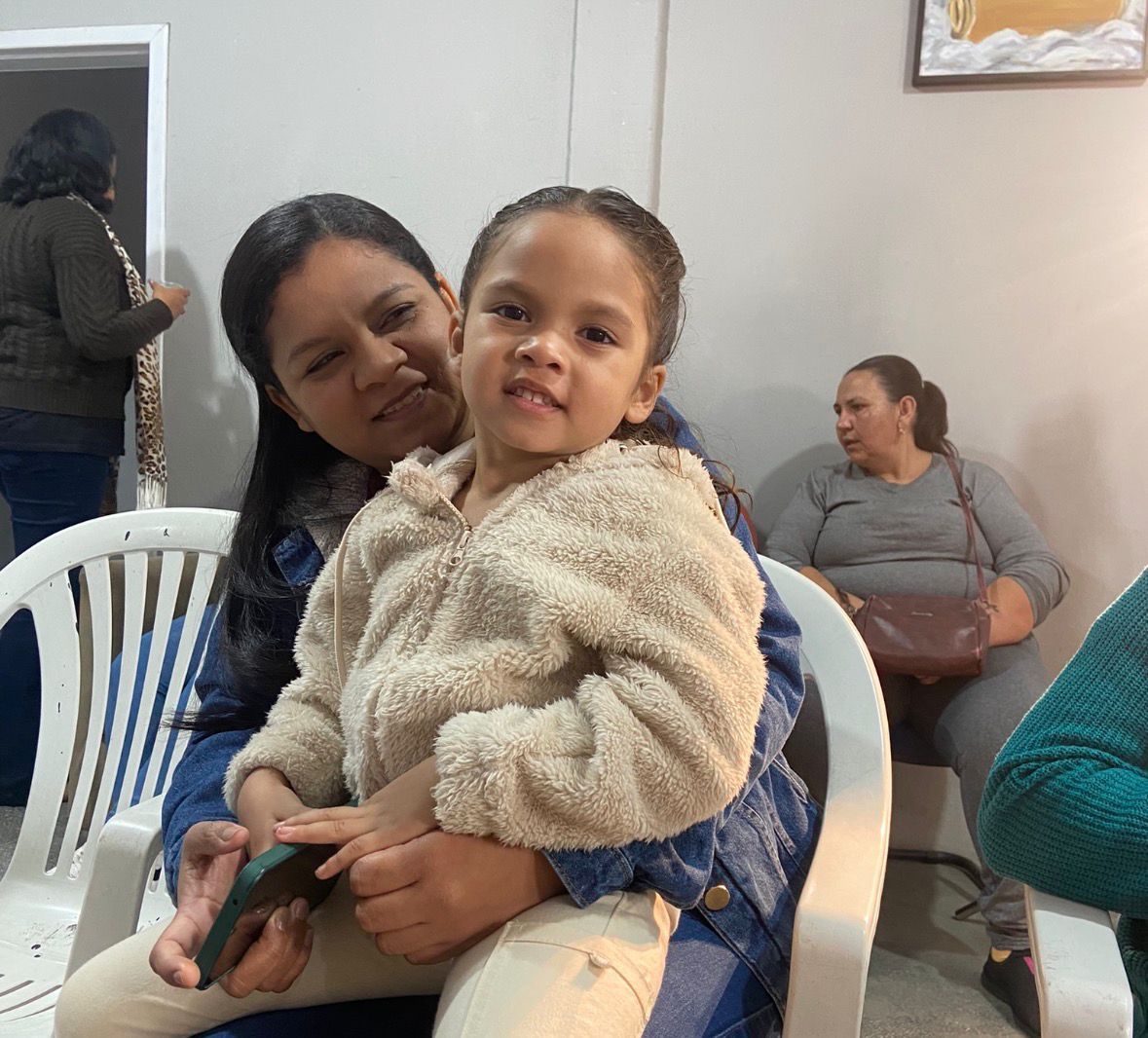 Uma criança sorridente, vestindo uma jaqueta felpuda, está sentada no colo de um adulto em um espaço interno, com outras pessoas sentadas por perto.