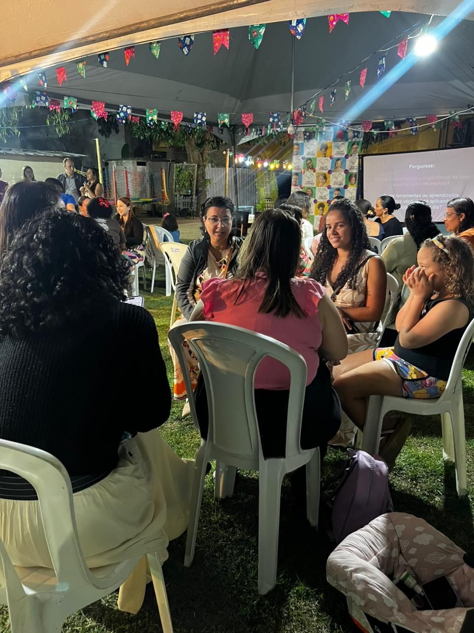 Pessoas sentam-se em cadeiras brancas em uma reunião ao ar livre à noite, decorada com bandeiras triangulares coloridas acima delas.