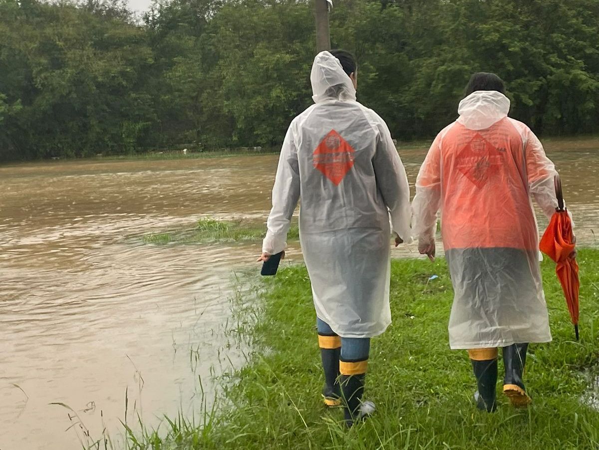 Duas pessoas com capas de chuva e botas caminham por uma área gramada inundada.