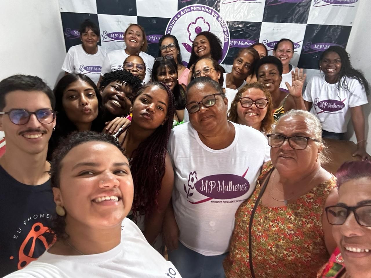 Um grupo de pessoas tirando uma selfie em frente a um fundo quadriculado em preto e branco com a marca